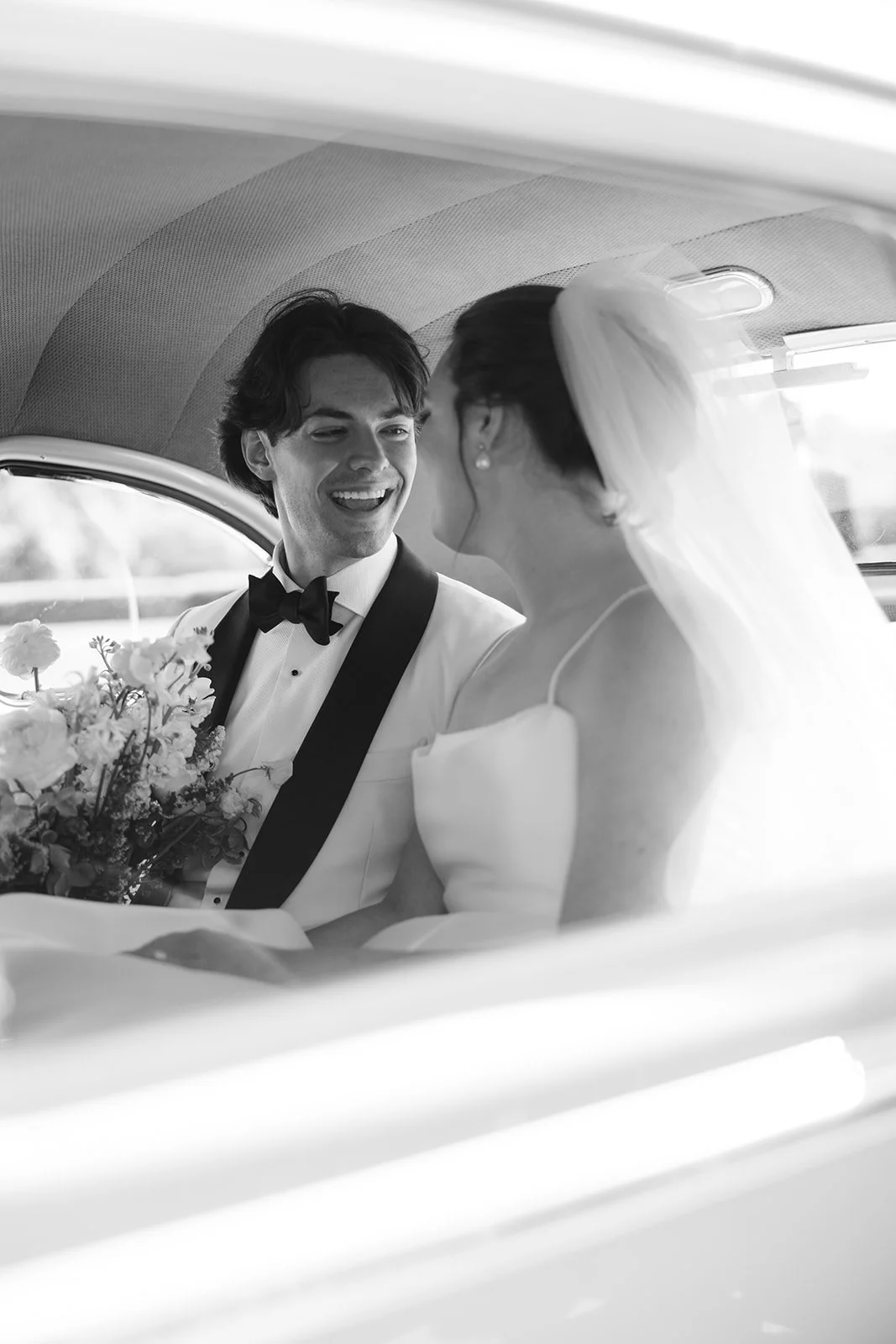 Black-and-white wedding photo of a smiling groom in a tuxedo and bride with a veil sitting together in a vintage car, holding flowers.