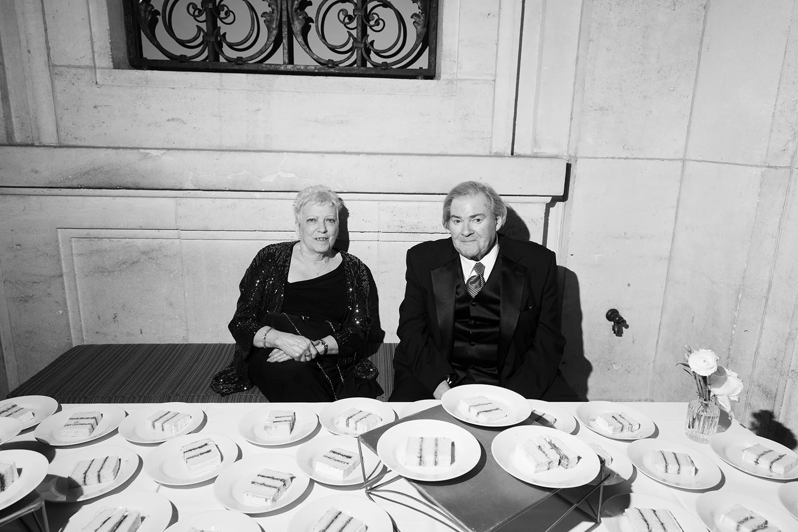 Black-and-white photo of an elegantly dressed couple seated behind a table of plated finger sandwiches at an event.