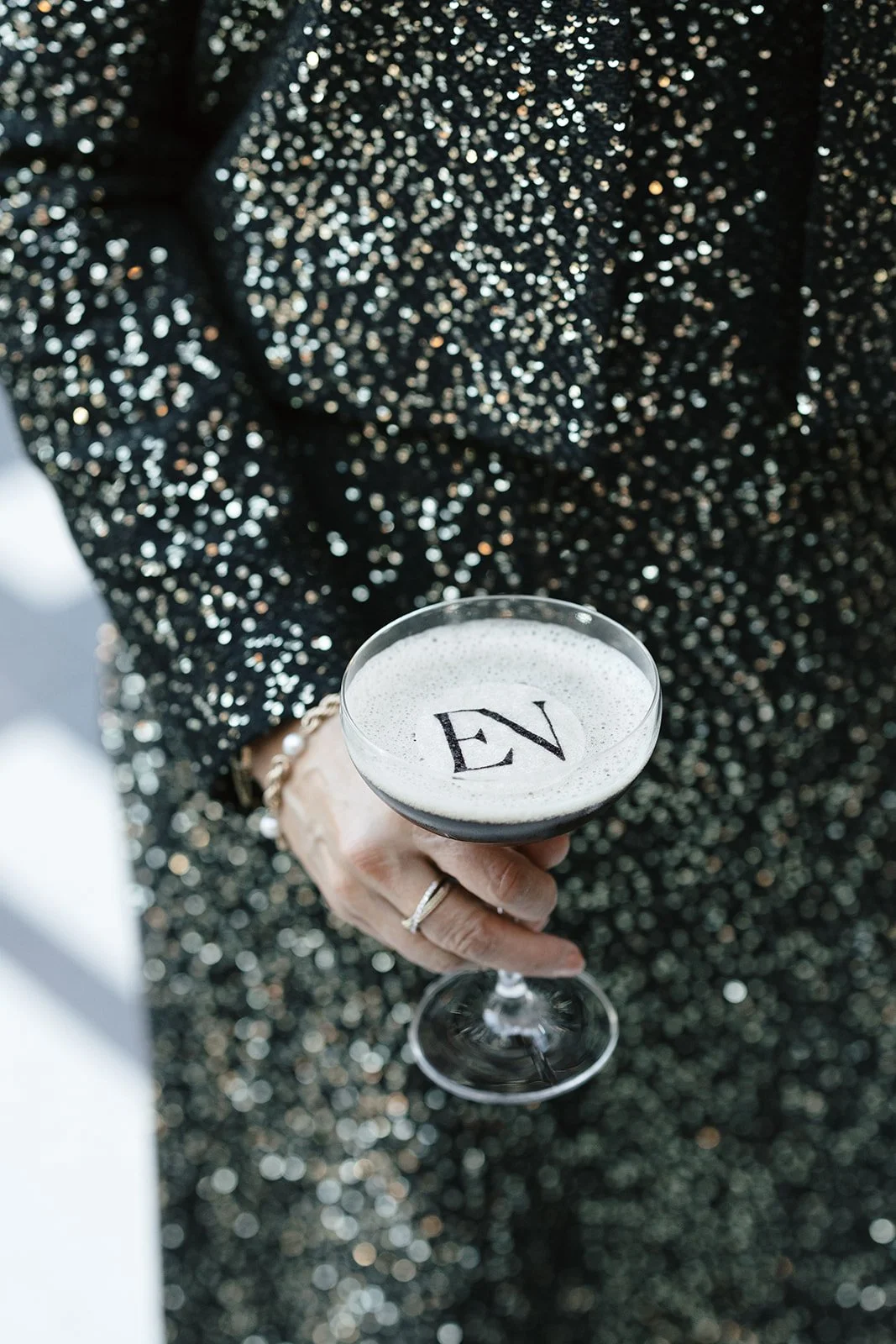 Hand holding a foamy cocktail in a coupe glass with an “EN” garnish against a black sequined background