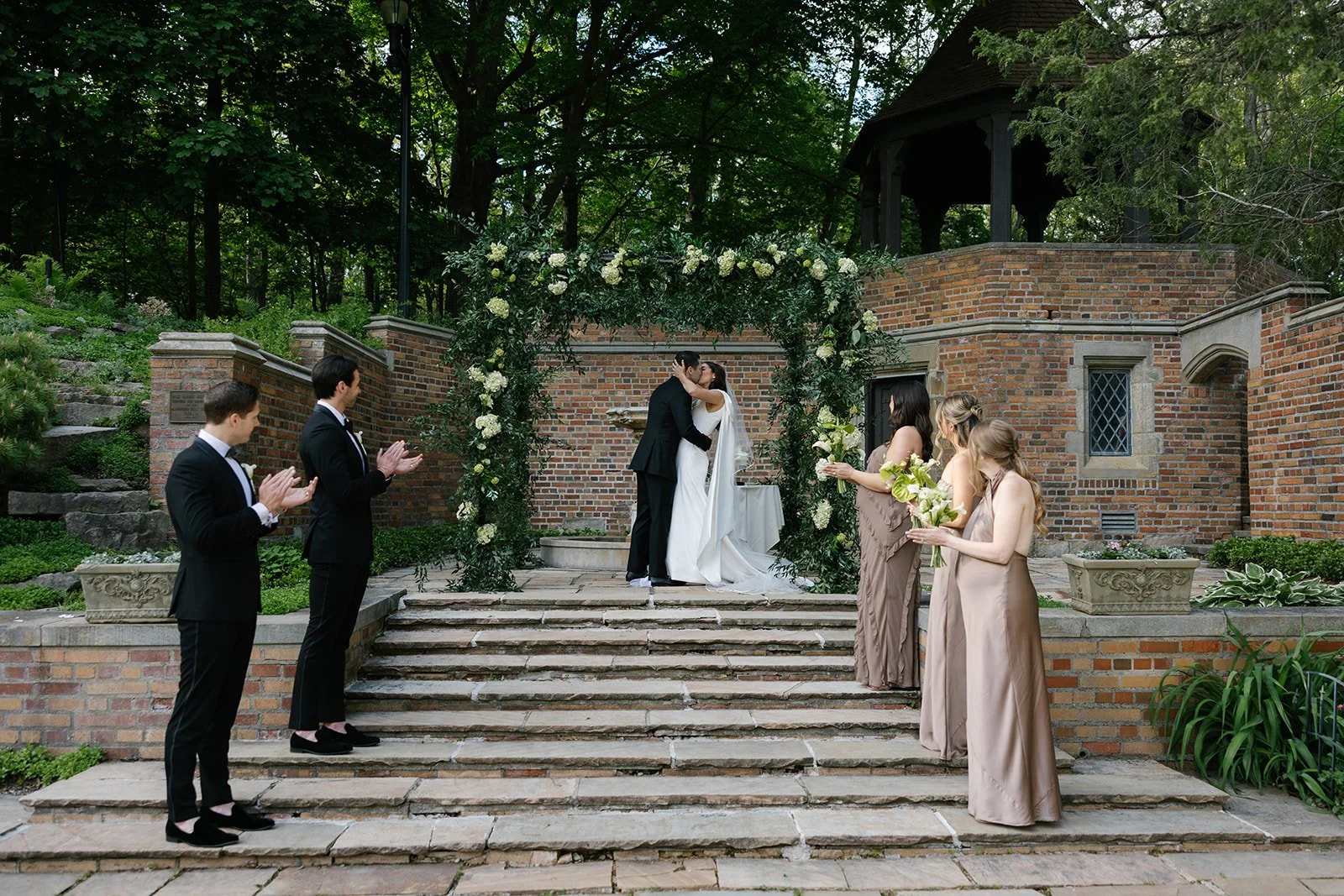 Bride and groom kissing beneath a floral wedding arch as bridesmaids and groomsmen applaud in a garden courtyard
