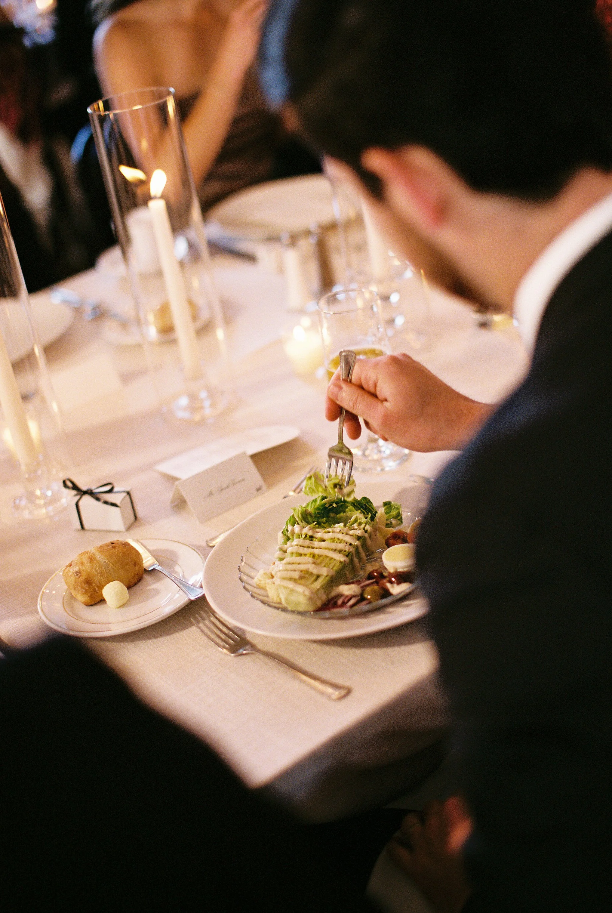 Elegant restaurant table with candles, wine glasses, bread, and a plated salad while a diner cuts food with a fork.
