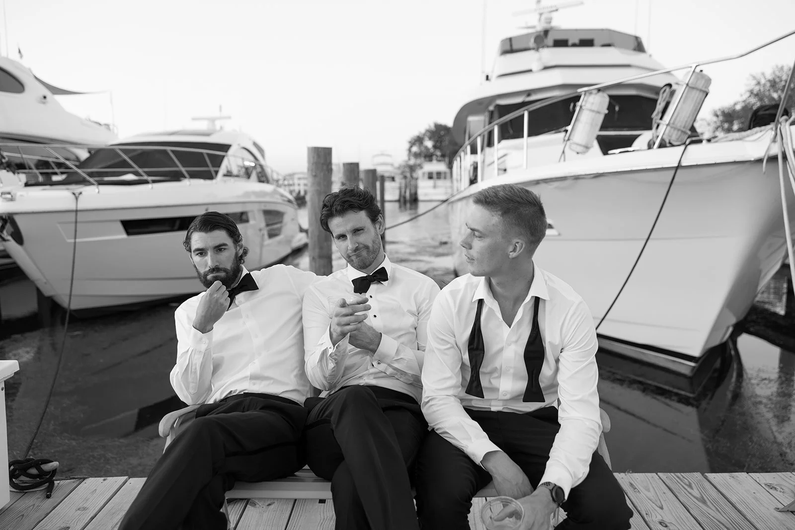 Three men in formal white shirts and bow ties sitting on a marina dock among luxury yachts