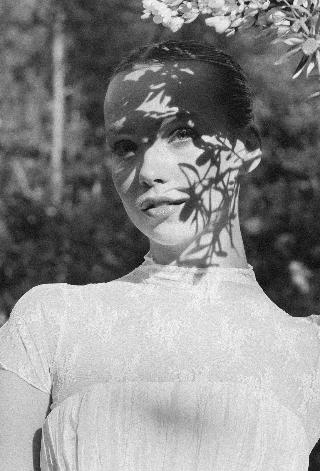 Black-and-white portrait of a woman in a white dress with leafy shadows cast across her face and neck