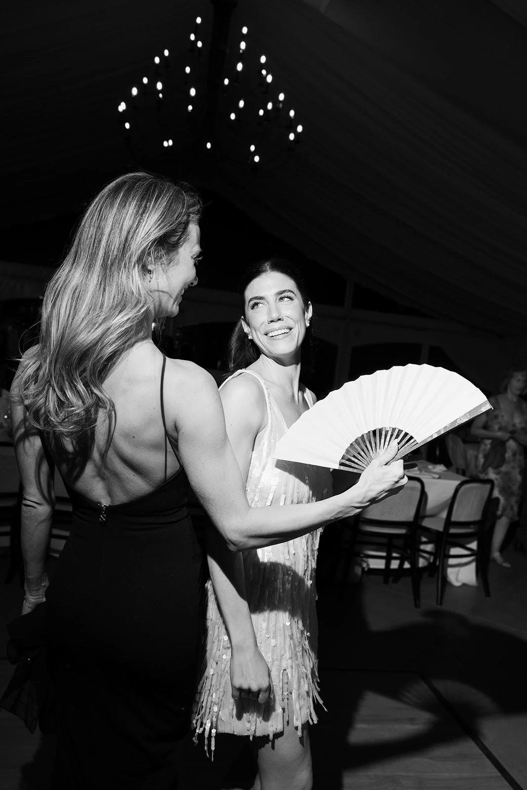 Black-and-white photo of two women at a party, with one holding an open folding hand fan beneath a chandelier.