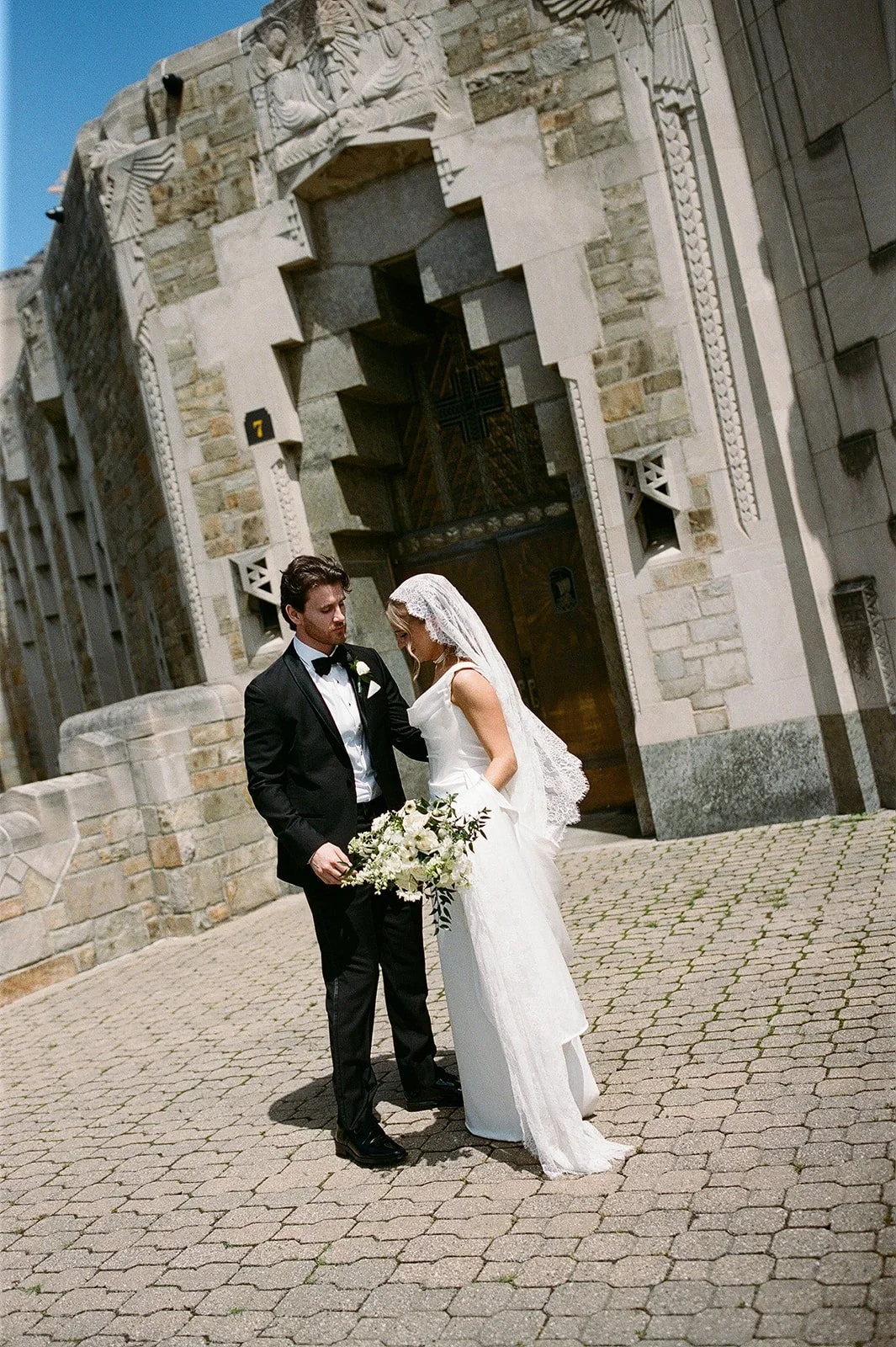 Bride and groom posing outside an ornate stone church, with the bride holding a white floral bouquet.