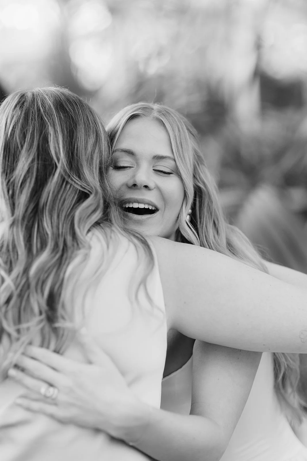 Black-and-white portrait of two women embracing outdoors, with one smiling and eyes closed.