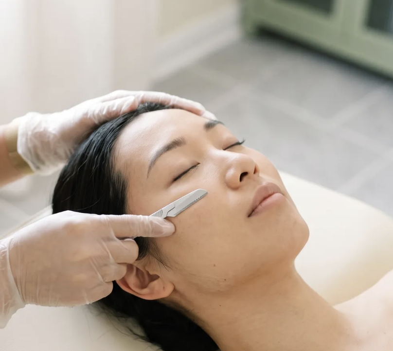 A woman receives a facial treatment in a spa or skincare clinic, lying down with a practitioner gently applying a tool to her face.