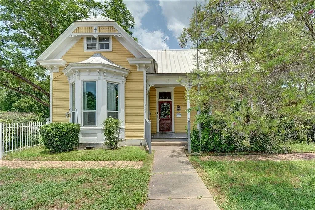Yellow Victorian-style house with white trim, a bay window, front porch, and greenery in the yard.