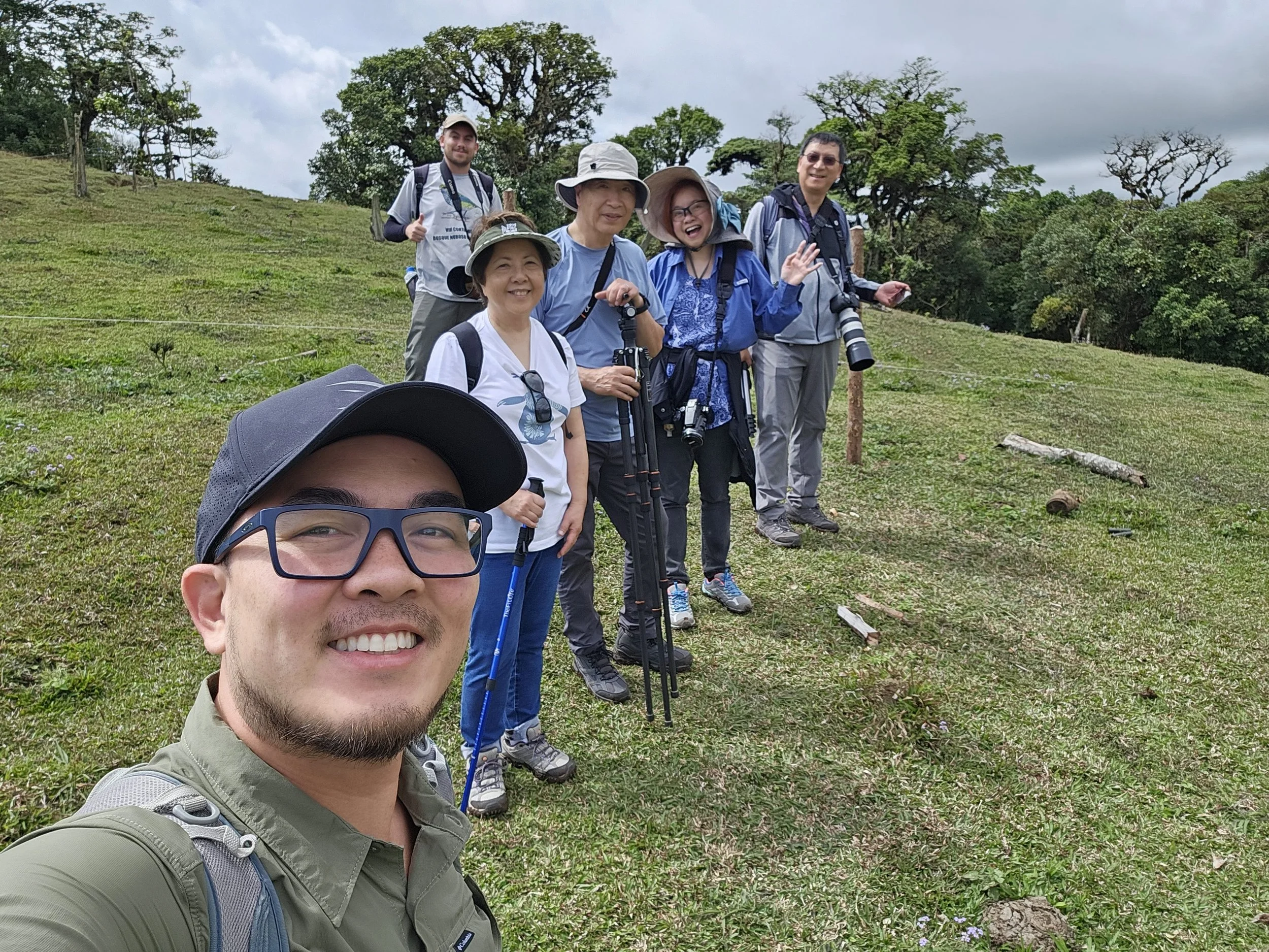 Group of six hikers taking a selfie on a grassy hillside with tropical trees and trekking poles.