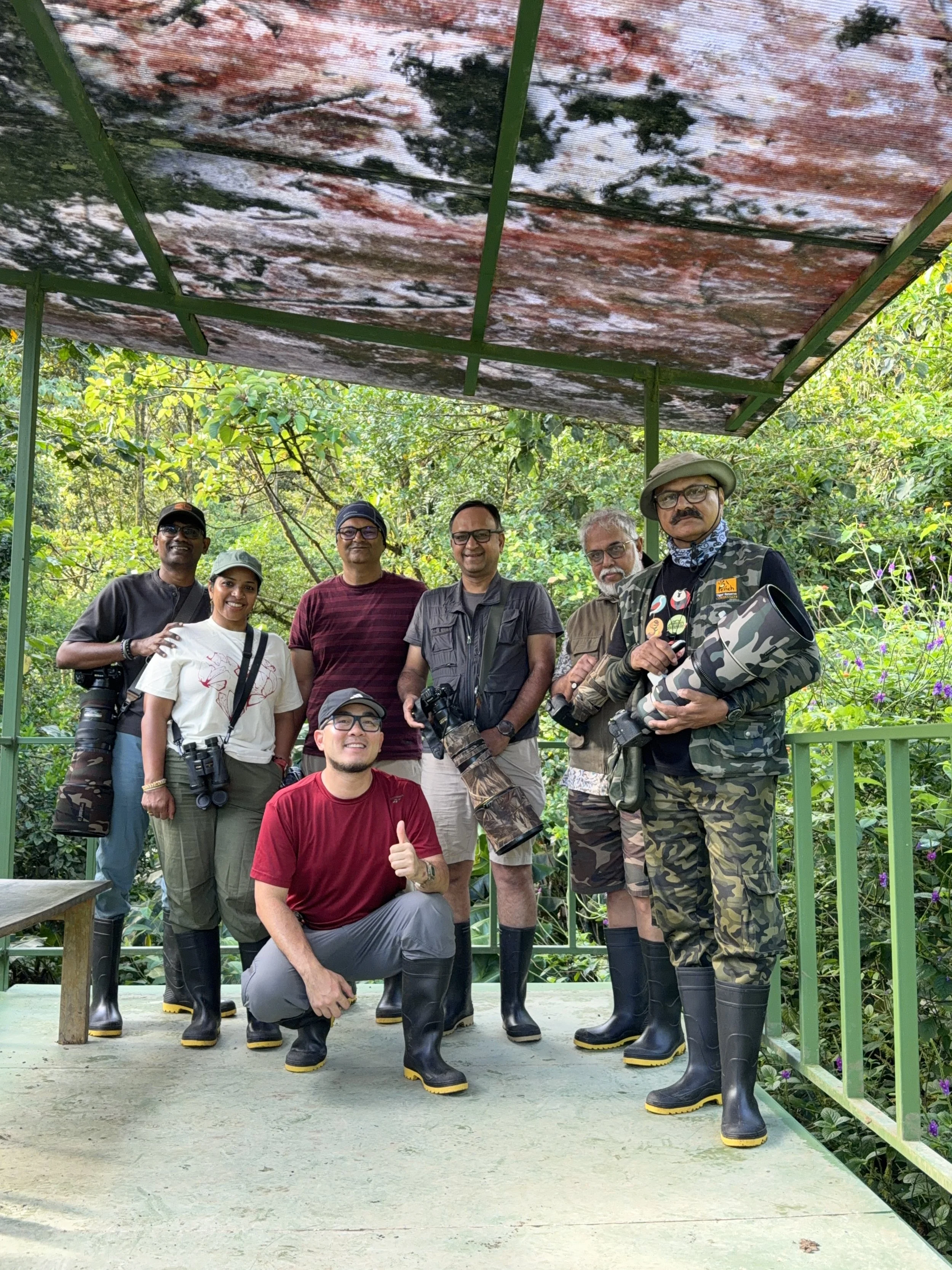 Group of photographers posing in a lush forest under a weathered shelter, wearing rubber boots and carrying cameras.