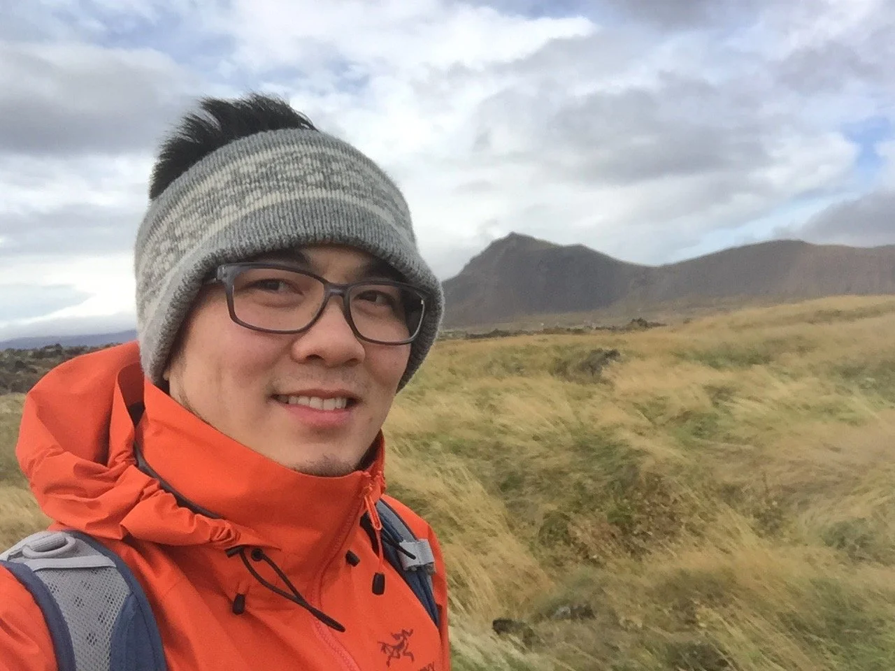 Hiker wearing an orange jacket, gray beanie, and glasses poses in a grassy mountain landscape under cloudy skies.