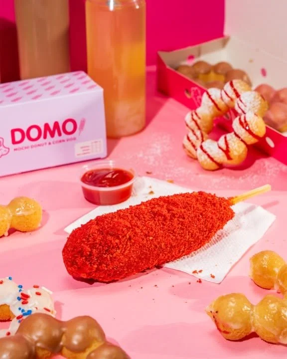 Fried corn dog with red seasoning on a paper napkin, surrounded by various fried foods and dipping sauces on a pink surface.