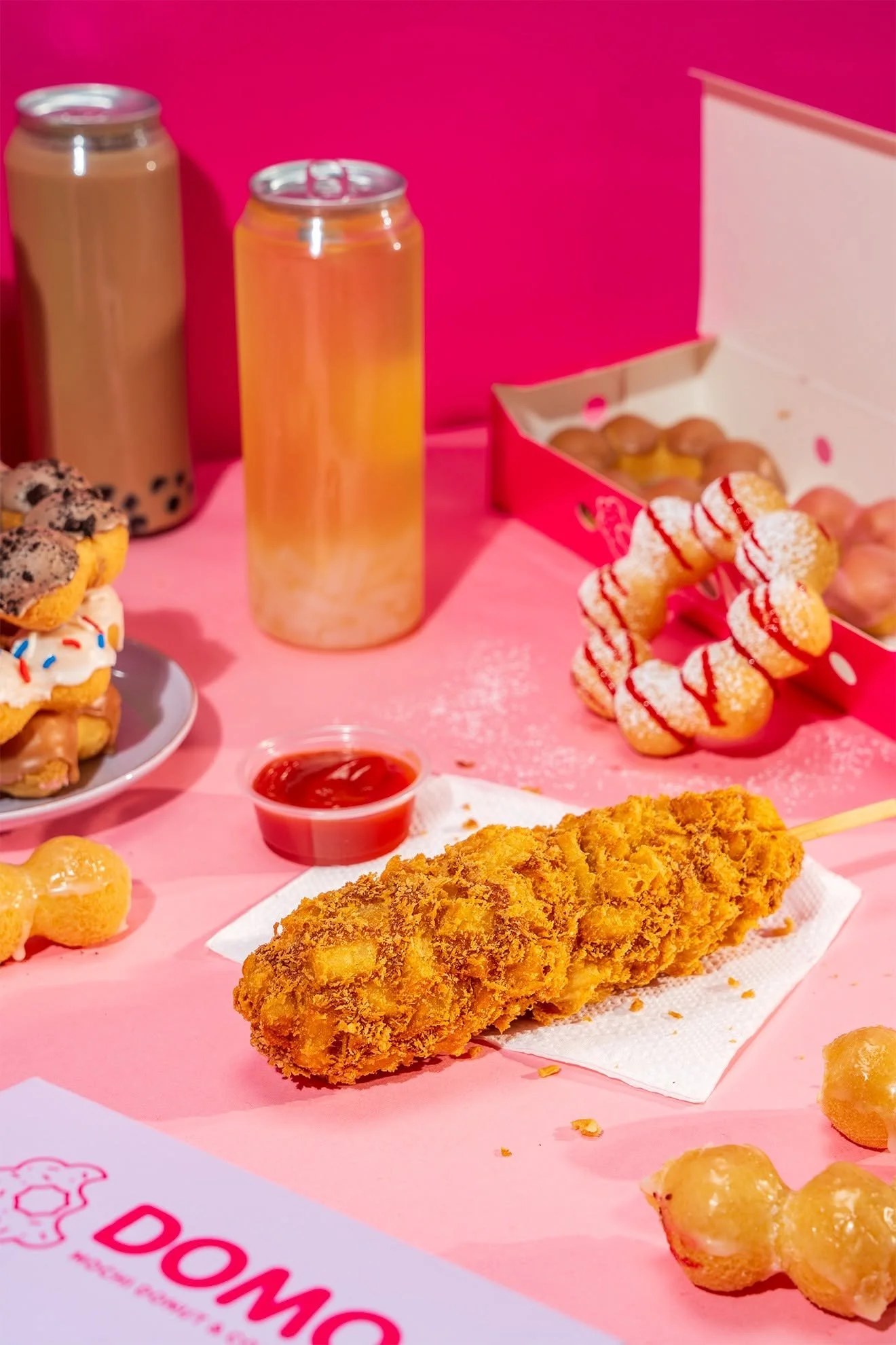 A spread of fried chicken, donuts, bubble tea, and assorted snacks on a pink table with a pink background.
