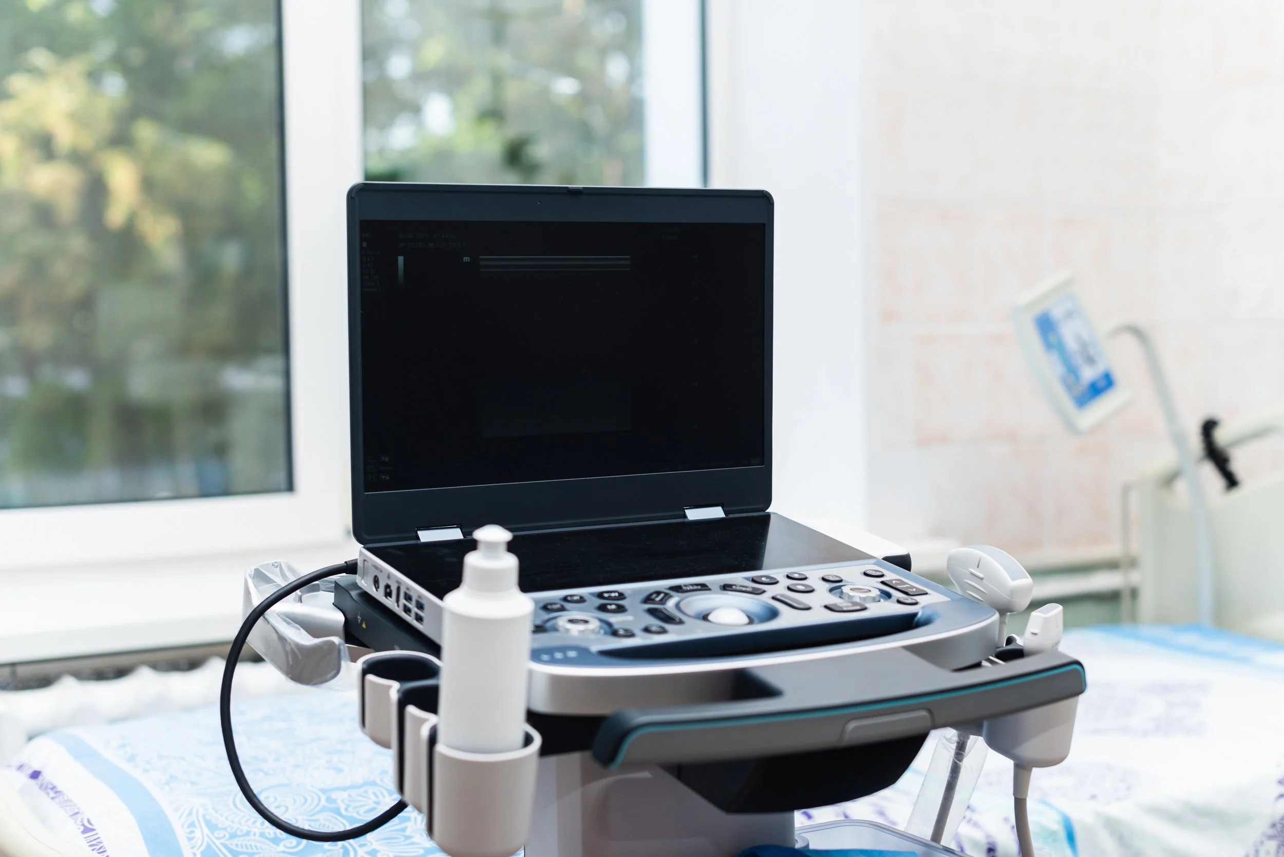 Medical ultrasound machine with monitor, probes, and controls in a clinical setting near a window with trees outside.