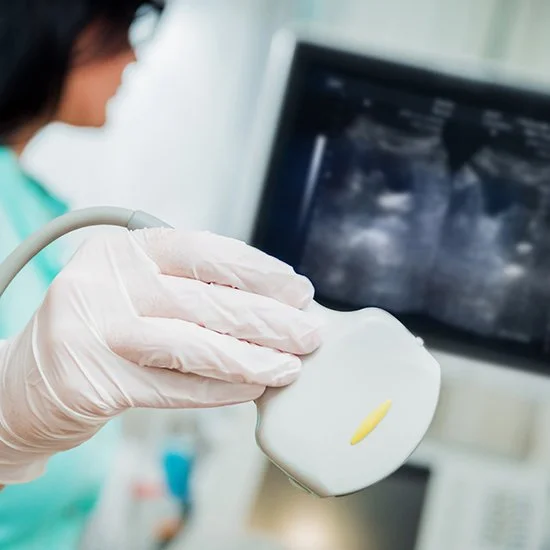 A healthcare professional wearing gloves holding an ultrasound probe in front of a monitor displaying a sonogram.