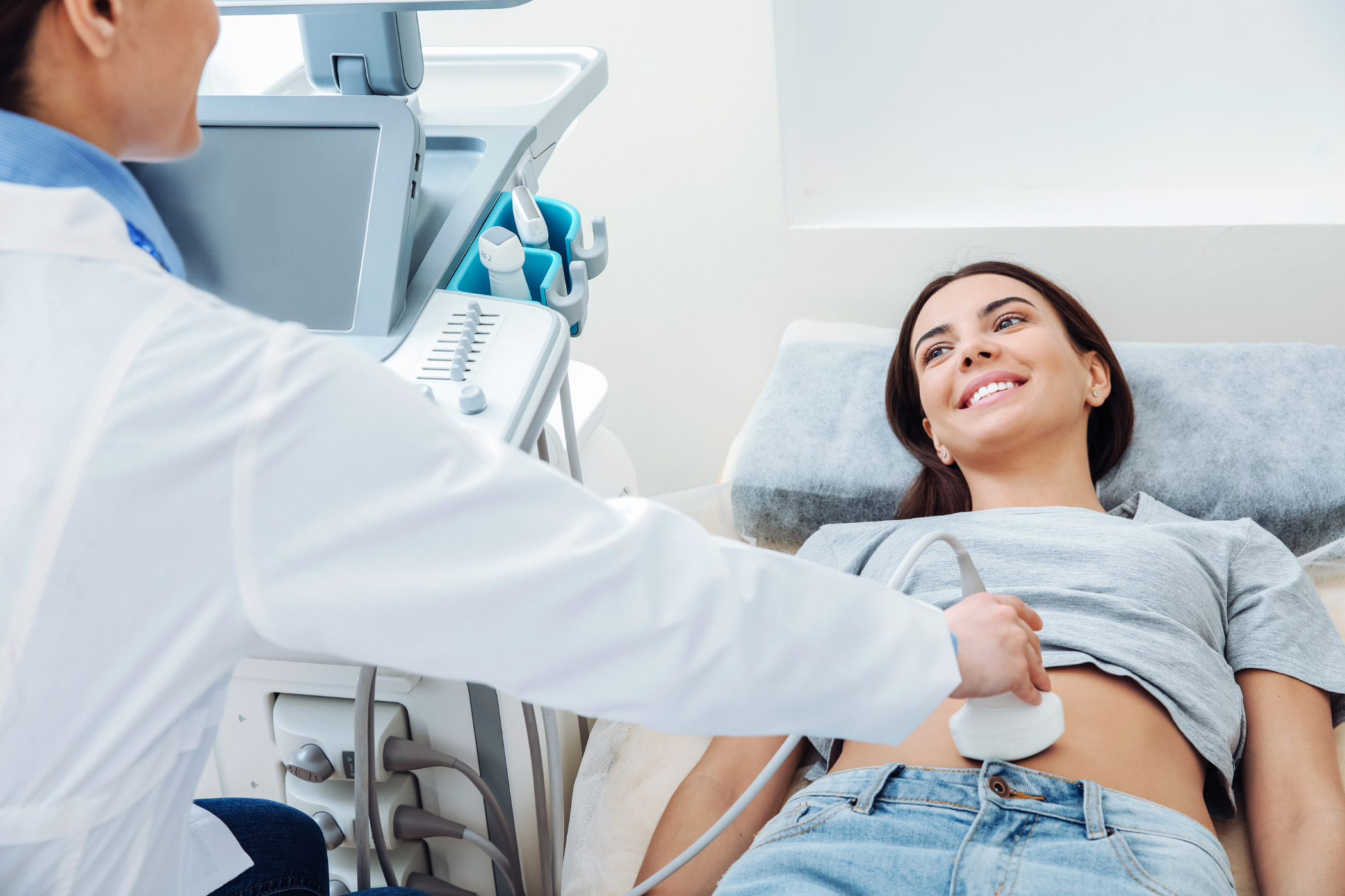 A woman lying on an examination bed at a medical clinic, smiling at the healthcare professional who is using an ultrasound device on her abdomen.