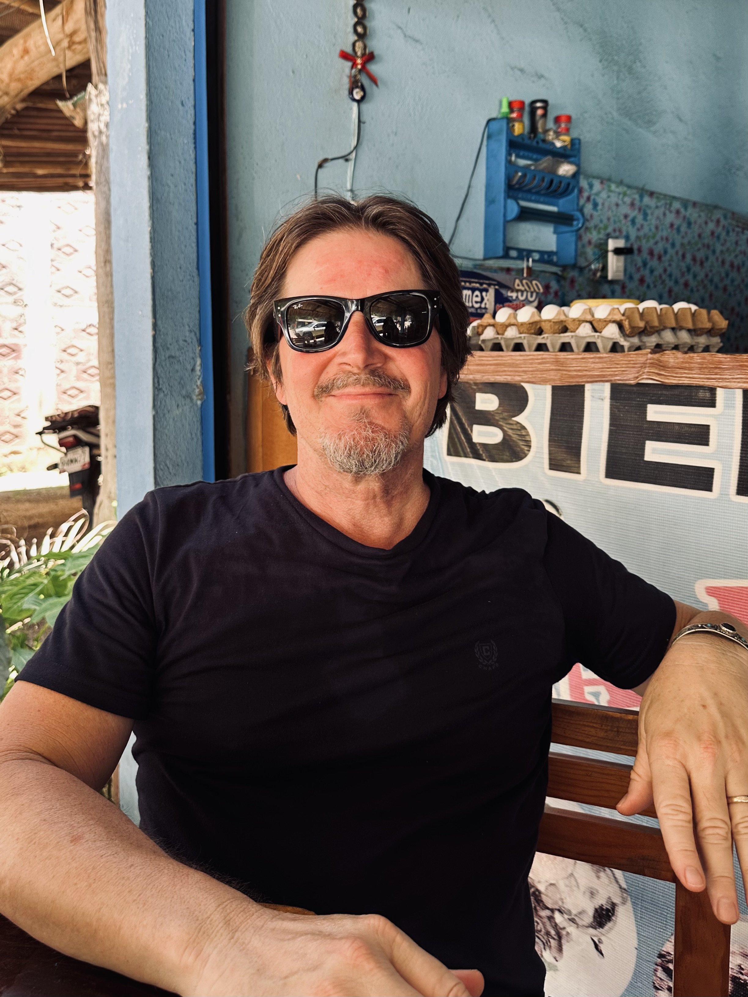 A middle-aged man with gray hair, beard, and mustache wearing sunglasses and a black t-shirt sitting outdoors, smiling at the camera. There are egg cartons and a sign in the background that reads 'BIE'.