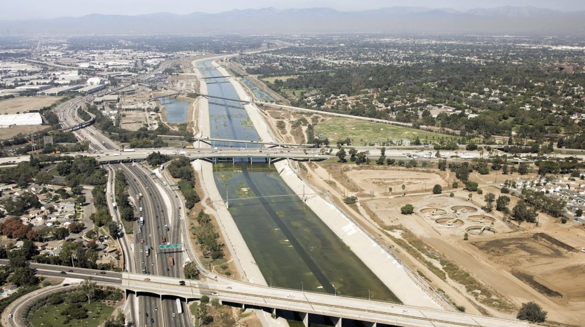 Aerial view of a city with highways, bridges, a waterway, residential area, and green spaces, with mountains in the background.