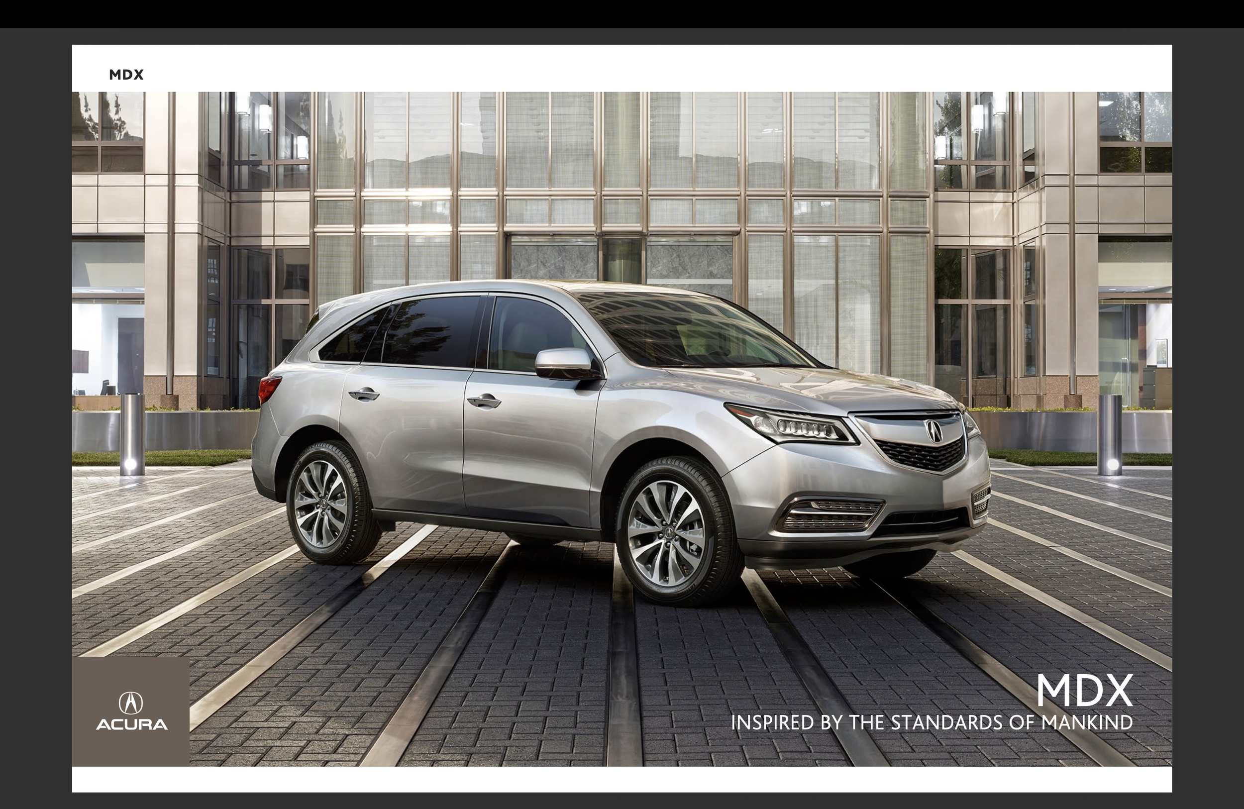 Silver Acura MDX SUV parked in a marked parking space in front of a modern glass building with mountains visible through the windows. The image includes branding and text indicating the MDX model and its inspiration from the standards of mankind.