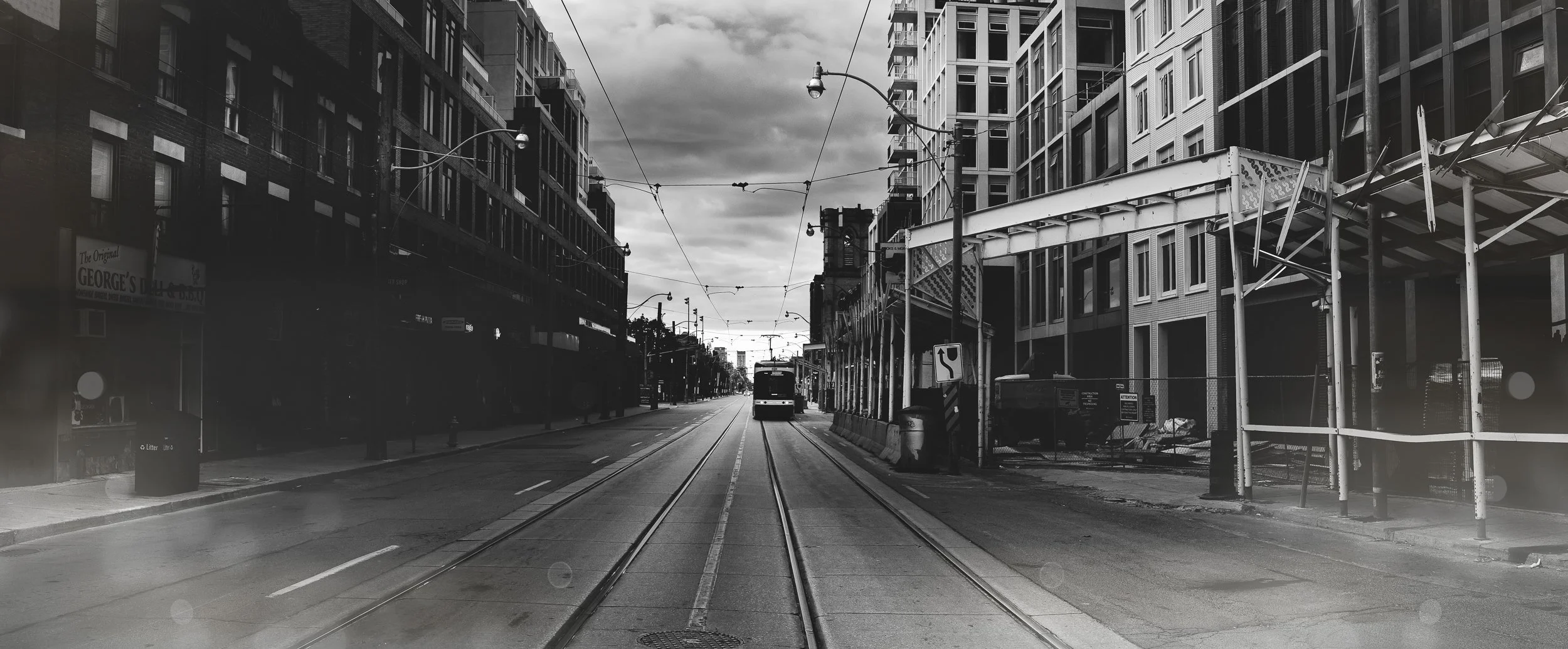 Black and white photo of city street with tram tracks, buildings on both sides, overhead wires, and a tram approaching in the distance.