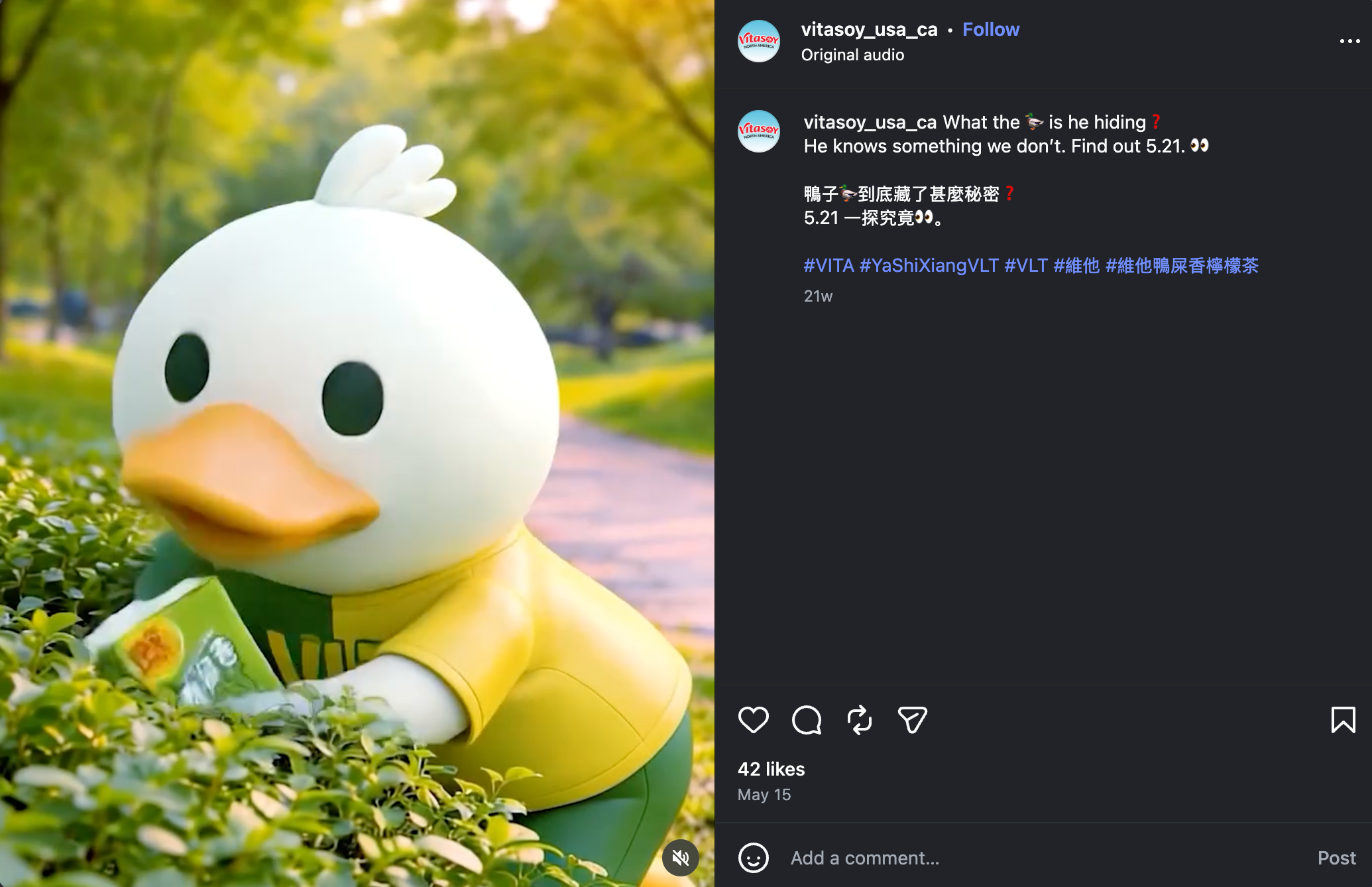A large white rubber duck with black eyes and an orange beak, wearing a yellow shirt, lying on green grass with trees in the background.