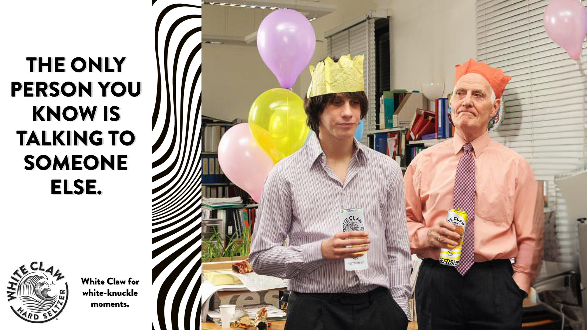 Two men at a celebration with balloons, wearing paper crowns, holding drinks, and standing in front of a bookshelf and a window with blinds.