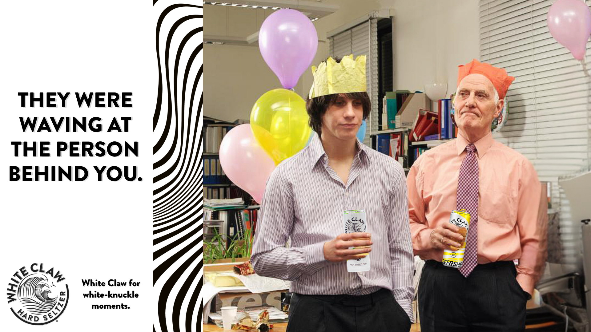 Two men at a celebration, both wearing paper hats and holding drinks, surrounded by colorful balloons and office bookshelves in the background.