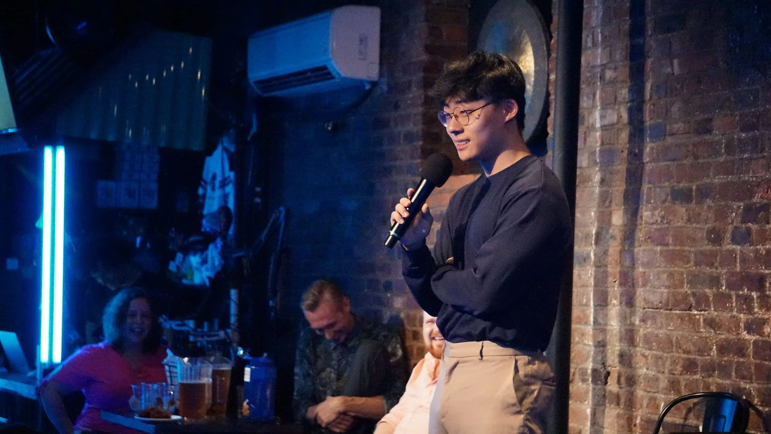 A young man with glasses holding a microphone and speaking to an audience at a bar or restaurant with brick walls and exposed pipes.