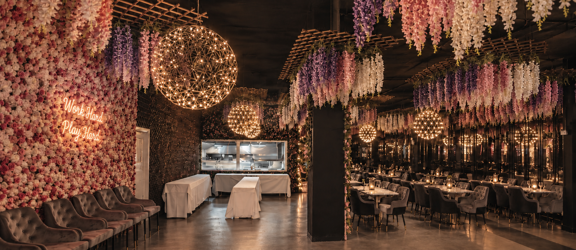 Interior of a decorated restaurant with floral walls, hanging flowers, illuminated spherical hanging lights, and tables set with candles.