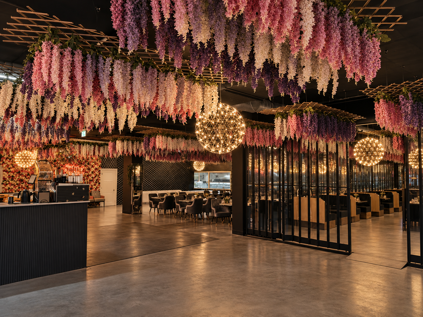 Interior of a restaurant or event space decorated with hanging pink, purple, and white flowers, star-shaped hanging lights, and glass partitions, with tables and chairs arranged inside.