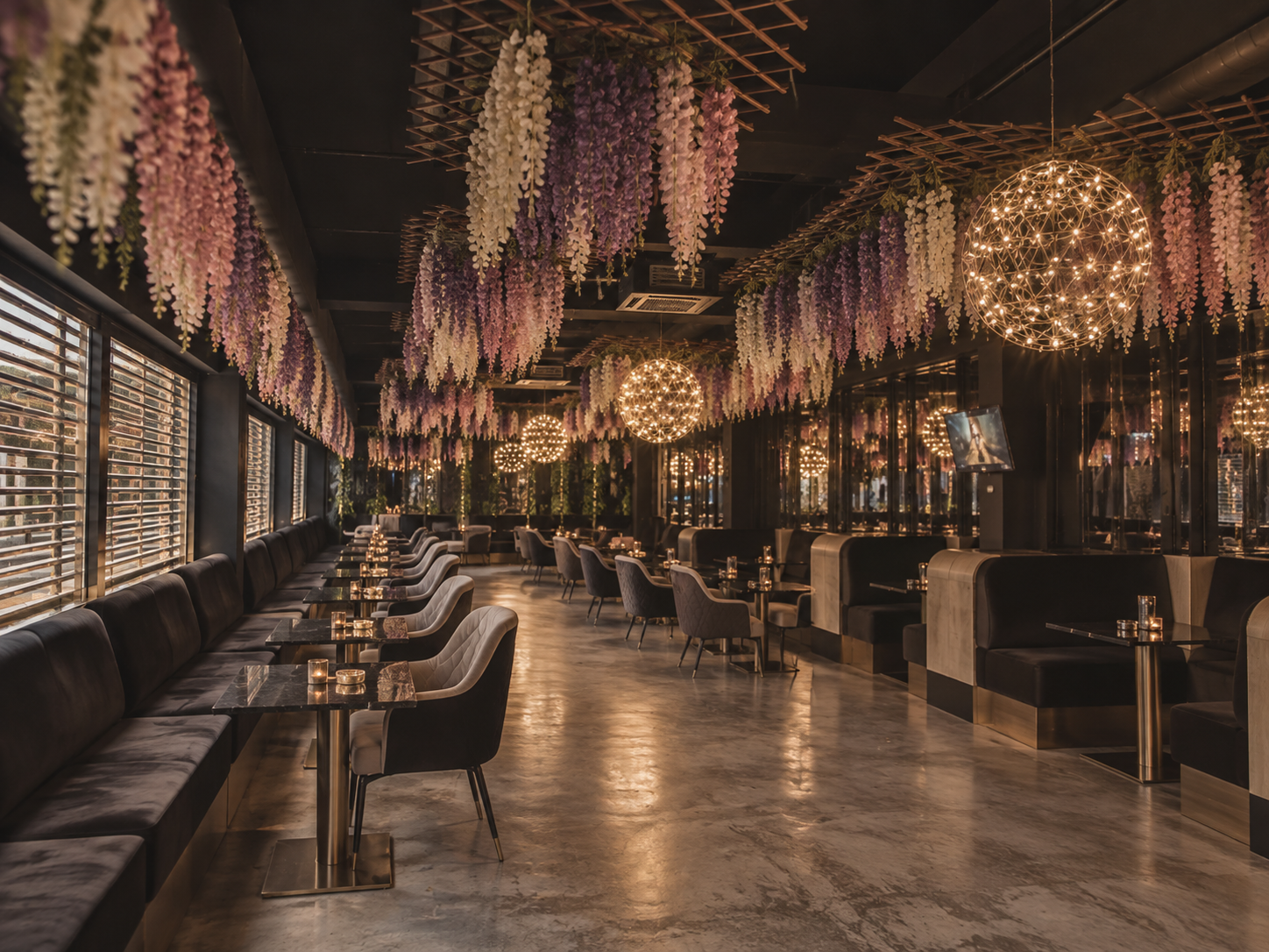 Interior of a dimly lit upscale restaurant or lounge with black walls and ceiling, decorated with hanging pink and purple floral arrangements and warm spherical chandeliers, featuring a row of black and beige upholstered booths and chairs along the windowed wall.