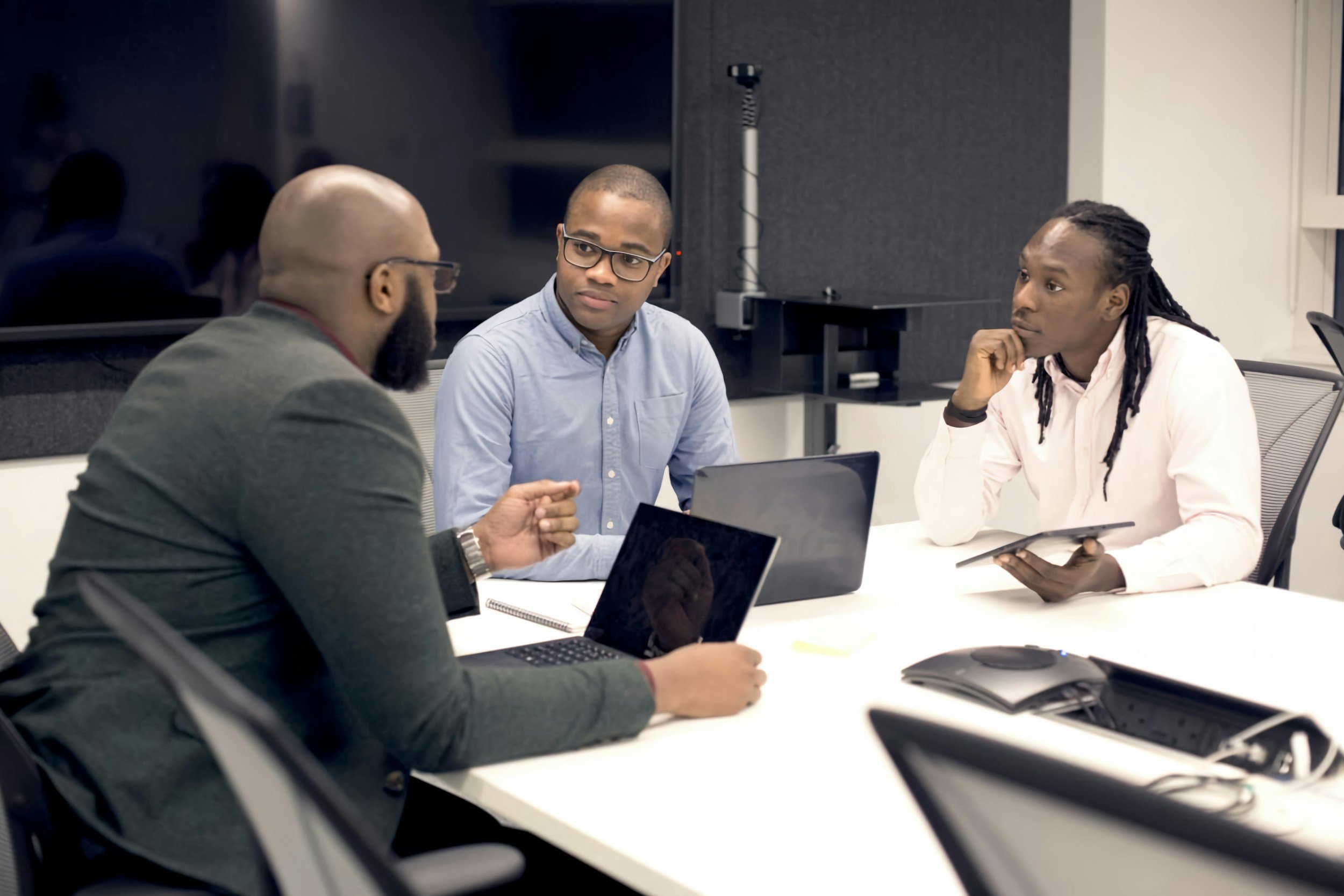 Three men in a professional meeting, two of them have laptops and one is holding a tablet, engaged in conversation in a conference room.