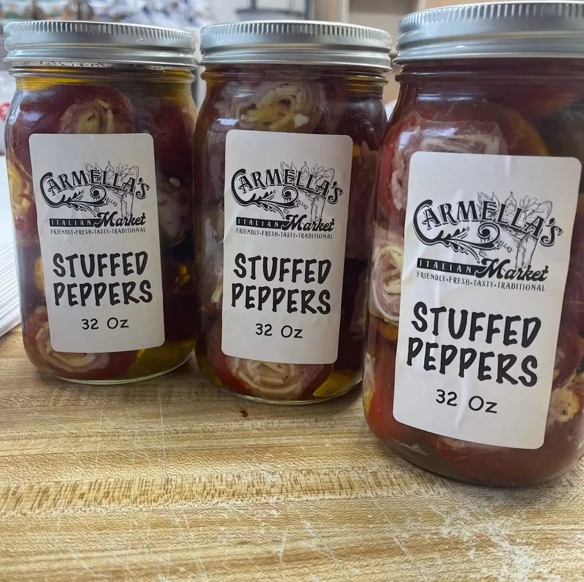Three glass jars labeled 'Armella's Italian Market Stuffed Peppers 32 Oz' on a wooden surface, containing stuffed peppers submerged in liquid.