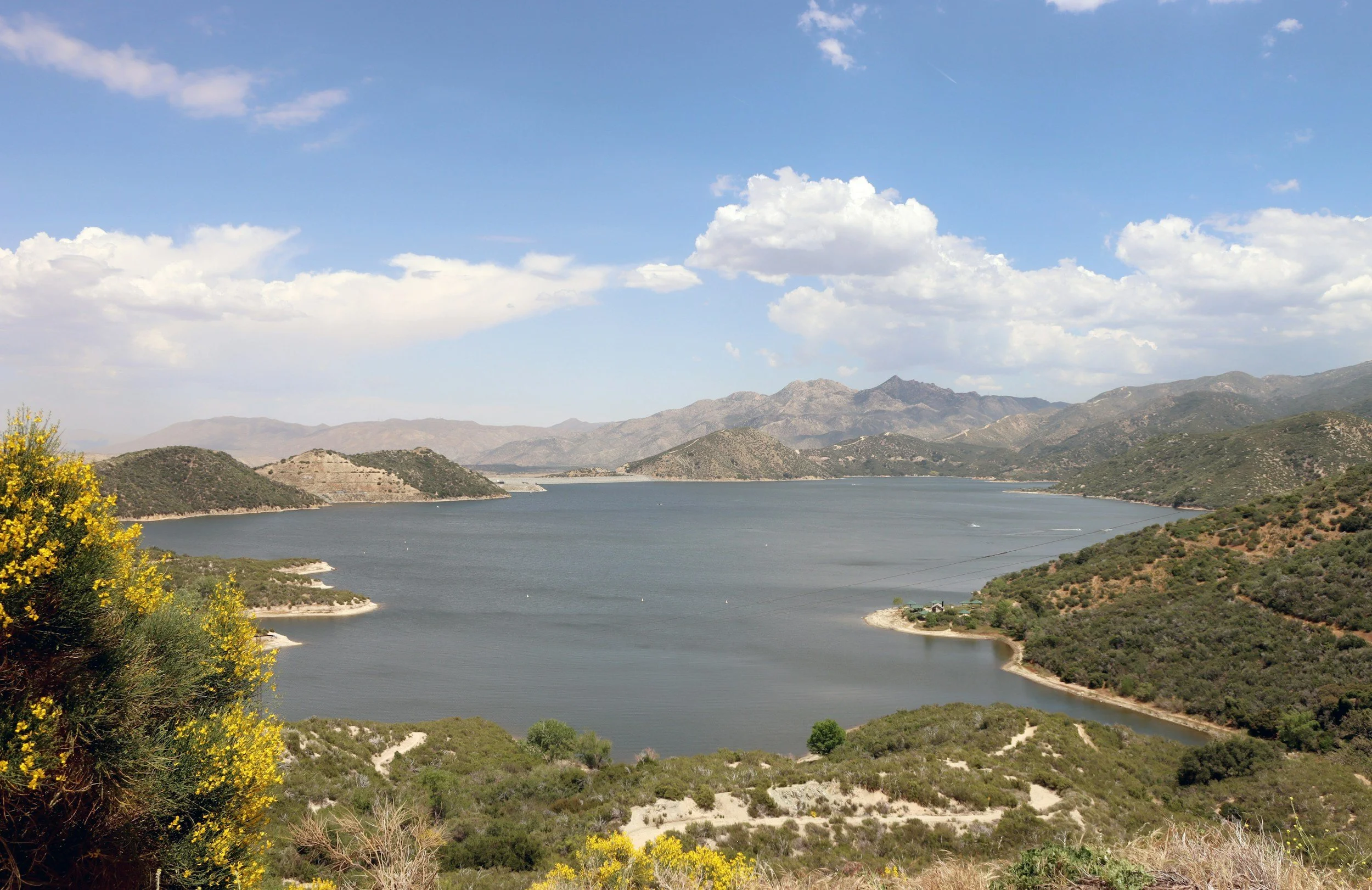 A large body of water, possibly a reservoir, surrounded by green hills and mountains under a partly cloudy sky with patches of blue.