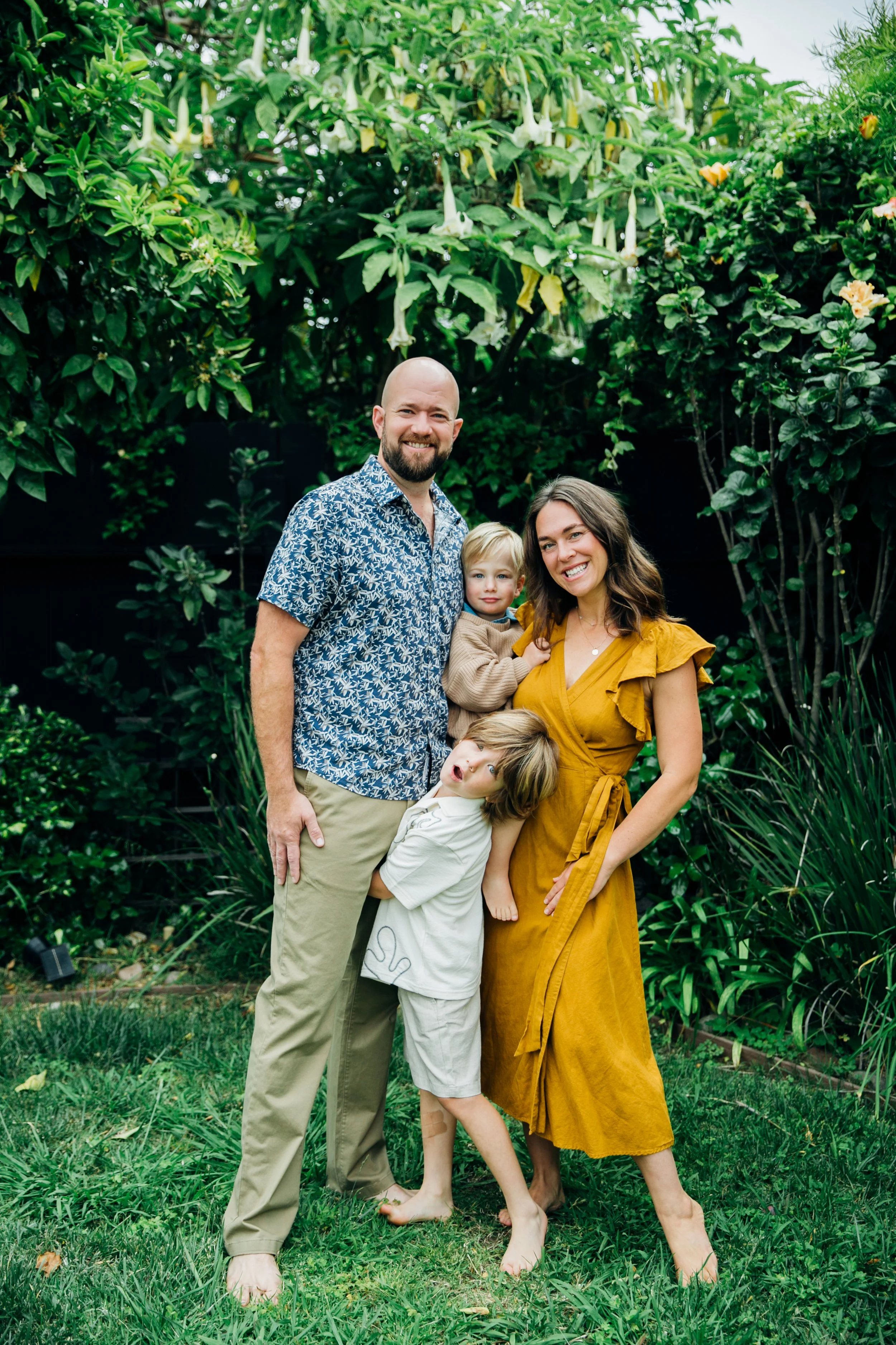 Family of four standing in a lush garden with green foliage, including large flowers hanging from a tree, smiling and relaxing outdoors.