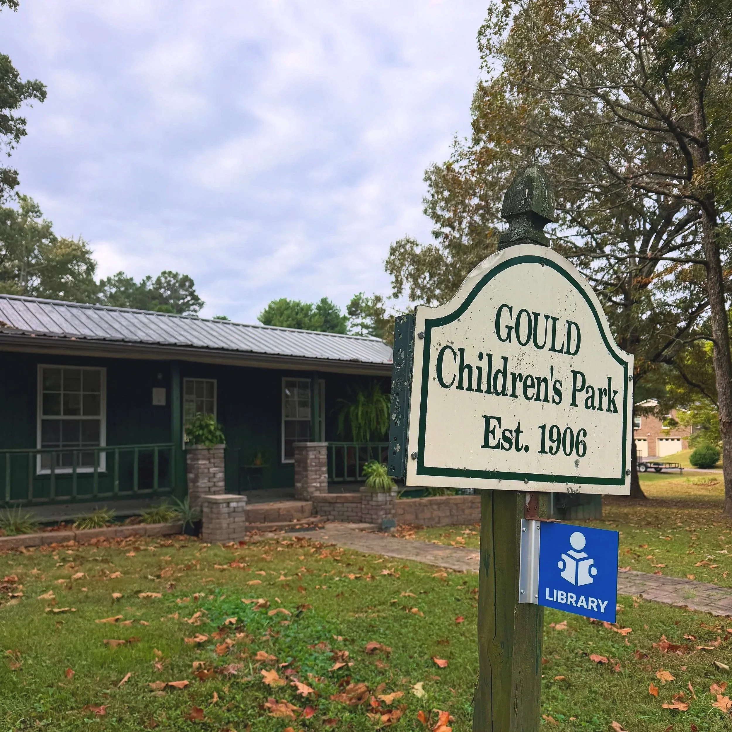 Sign for Gould Children’s Park, established in 1906, beside a small library building and trees.