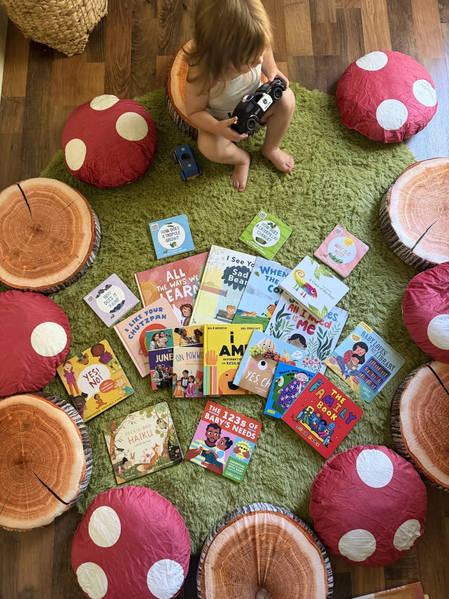 Child holding a toy police car on a green rug surrounded by children’s books, red mushroom cushions, and tree-stump seats.