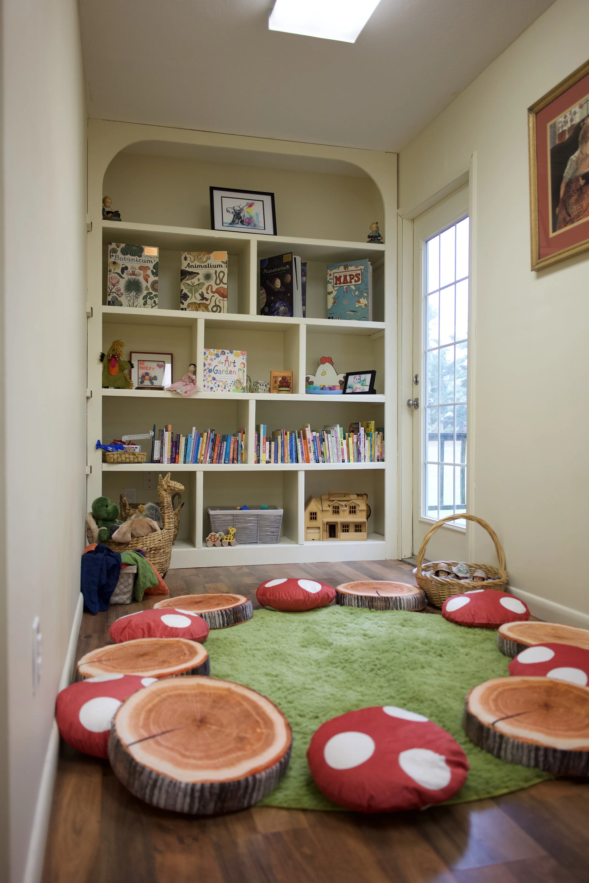 Children’s playroom with a bookshelf of books and toys, green rug, and red mushroom and tree-stump cushions.