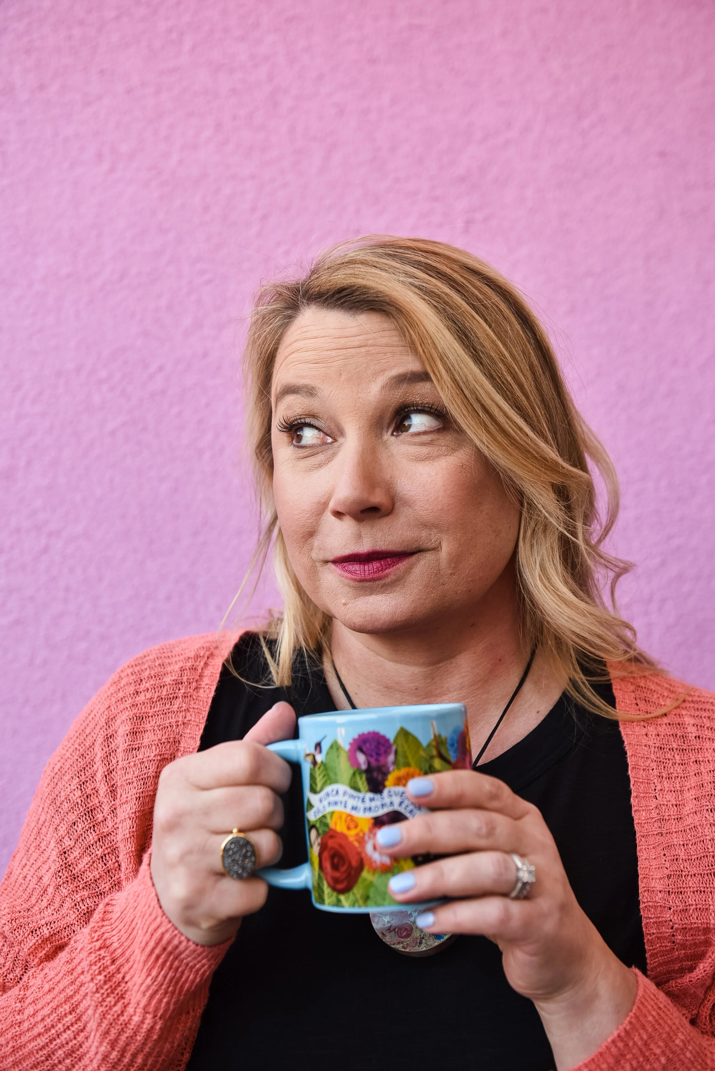 A woman with blonde hair holding a colorful floral mug, wearing a black top and a pink cardigan, against a pink wall background.