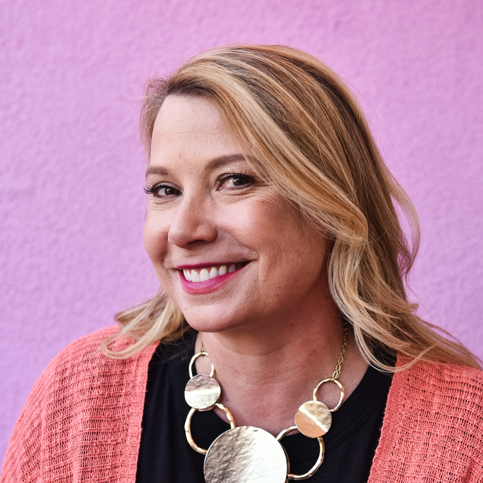 Close-up portrait of a smiling woman with wavy blonde hair, wearing a black top, an orange cardigan, and a large statement necklace, against a purple background.