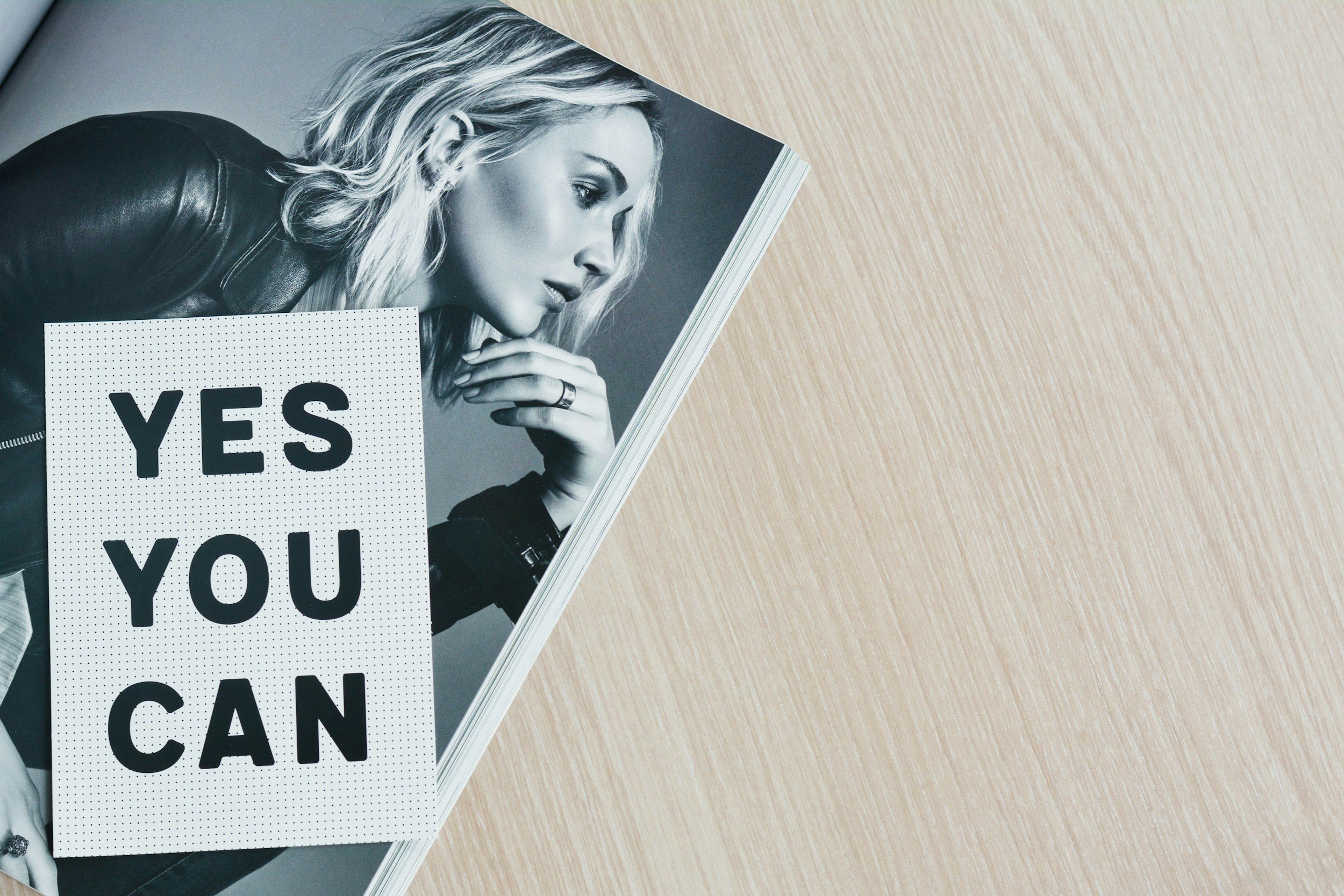 Black and white magazine page featuring a woman with wavy blonde hair and a leather jacket, lying on a wooden surface. A sign with bold text reads 'YES YOU CAN' partially covering her.