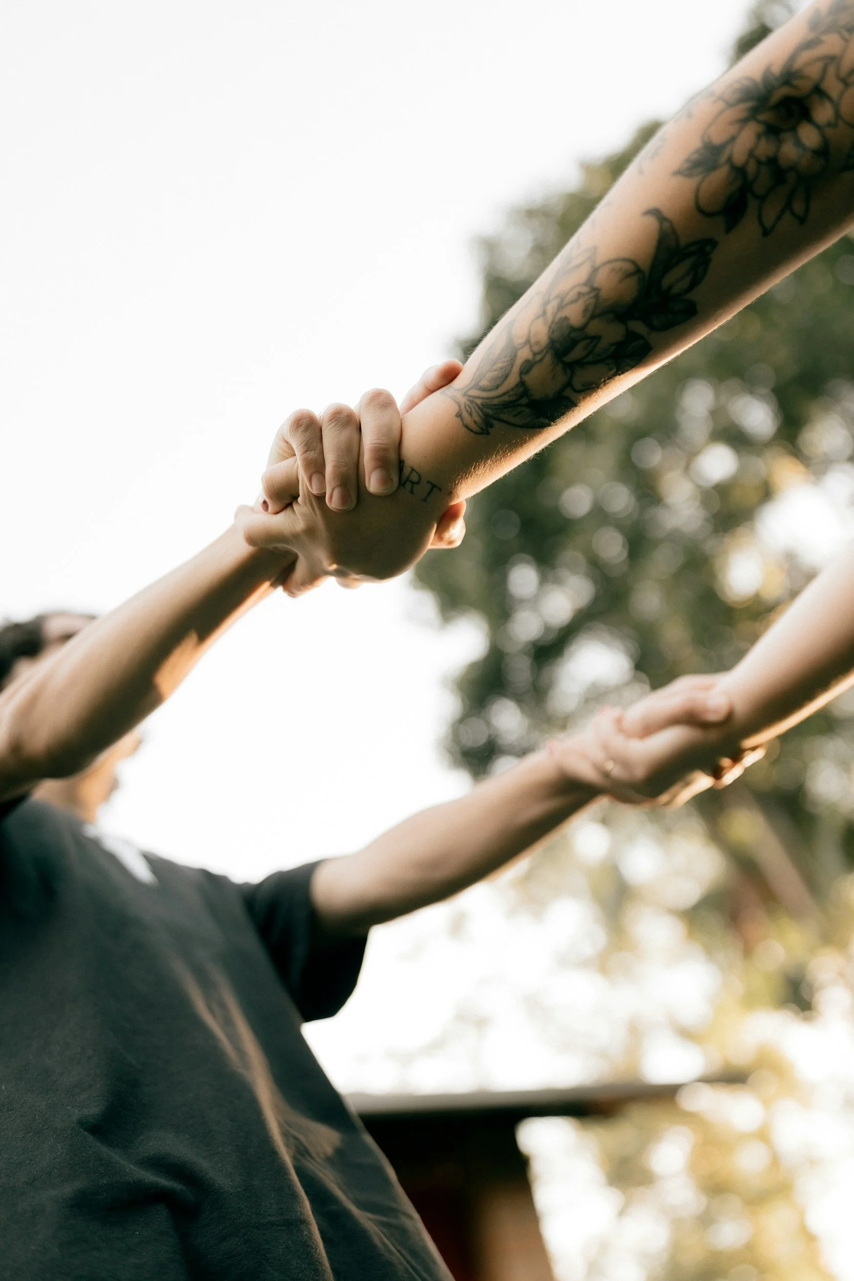 Multiple individuals holding hands, with one person's arm displaying tattoos, including flowers and the word 'RISE', against a background of blurred trees and sunlight.