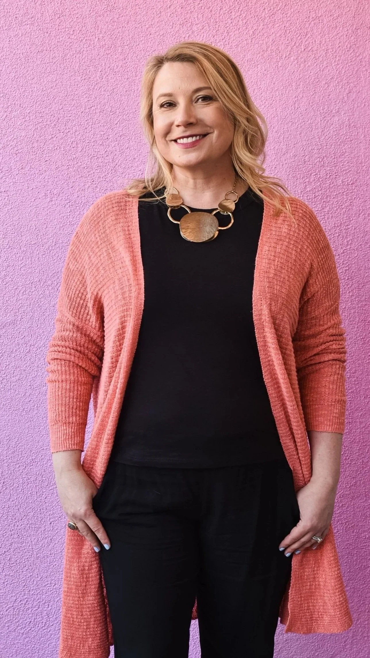 A woman with blonde hair is smiling and standing against a pink textured wall. She is wearing a black top, a coral cardigan, and a large statement necklace with a circular pendant. She has rings on her fingers and painted nails.
