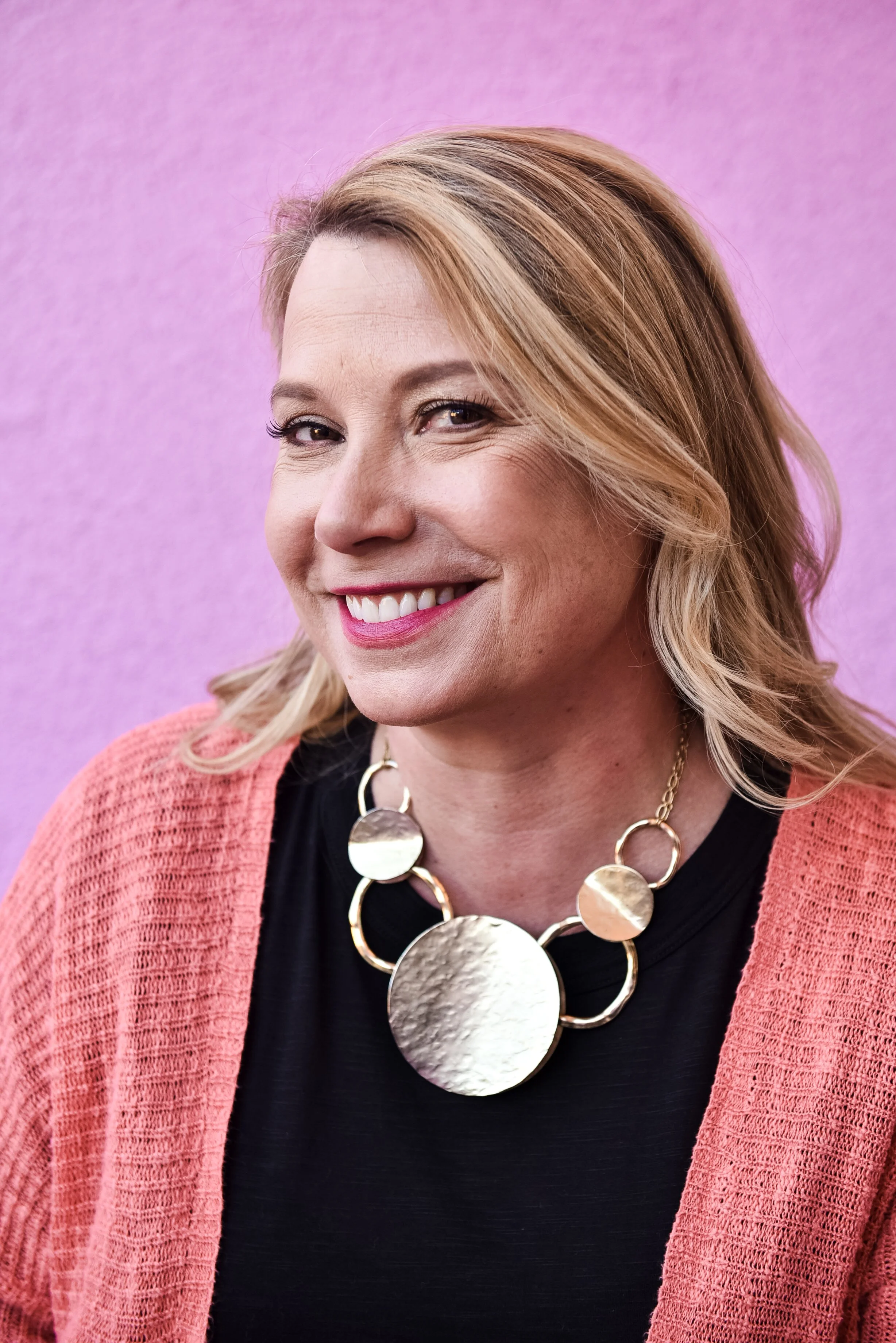 A woman with blonde hair smiling, wearing a black top, a pink cardigan, and a large statement necklace with circular gold and hammered metallic discs, in front of a purple background.