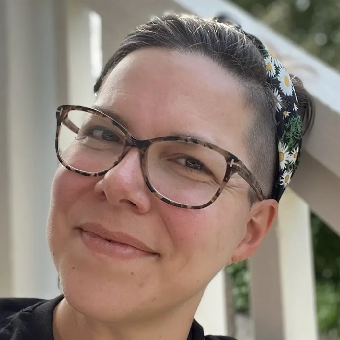 A person with short dark hair, wearing glasses and a floral headband, smiling outdoors.