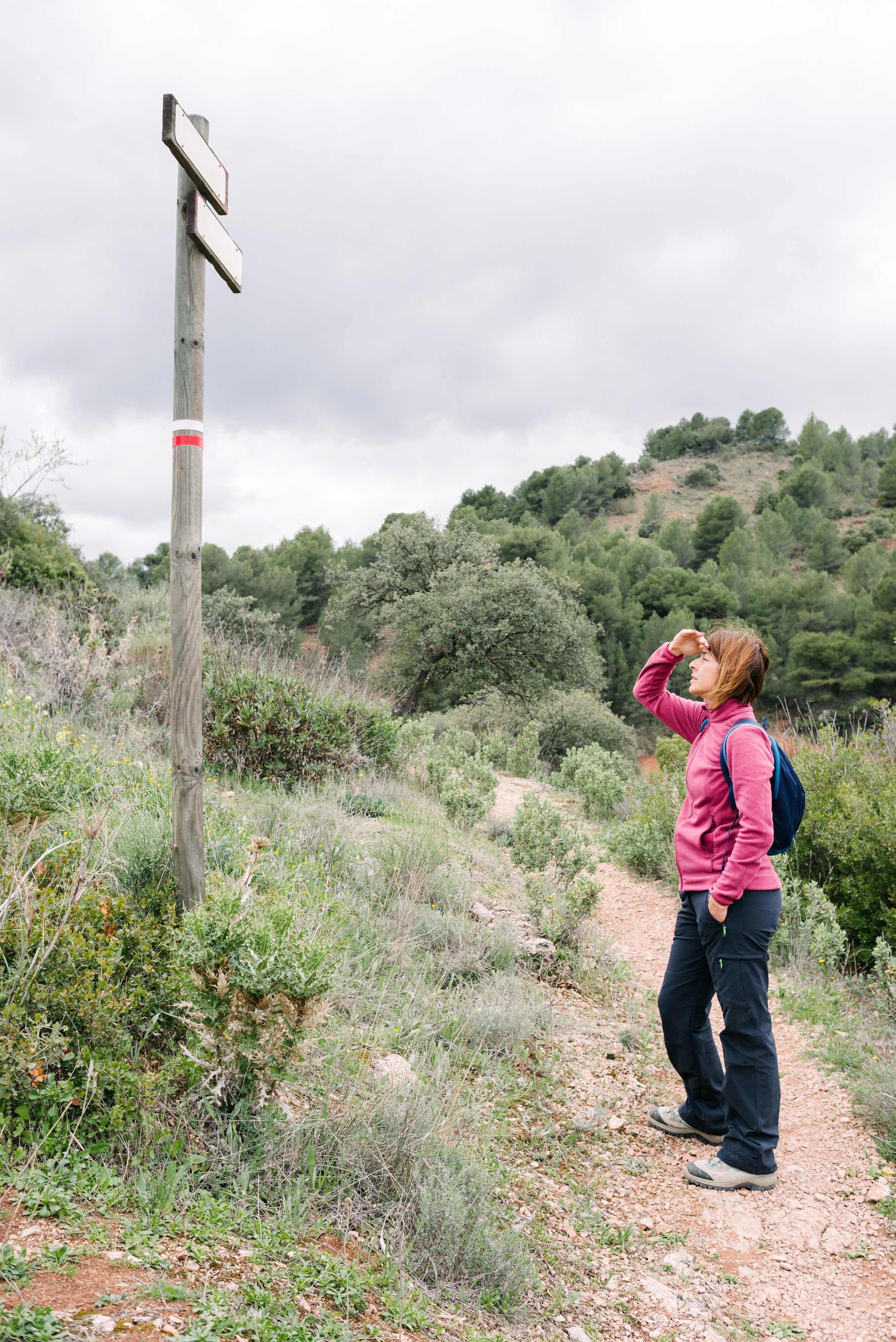 A woman with a backpack, dressed in a pink jacket and dark pants, standing on a dirt trail, holding her hand to her forehead while looking at a signpost in a hilly, wooded area with overcast skies.