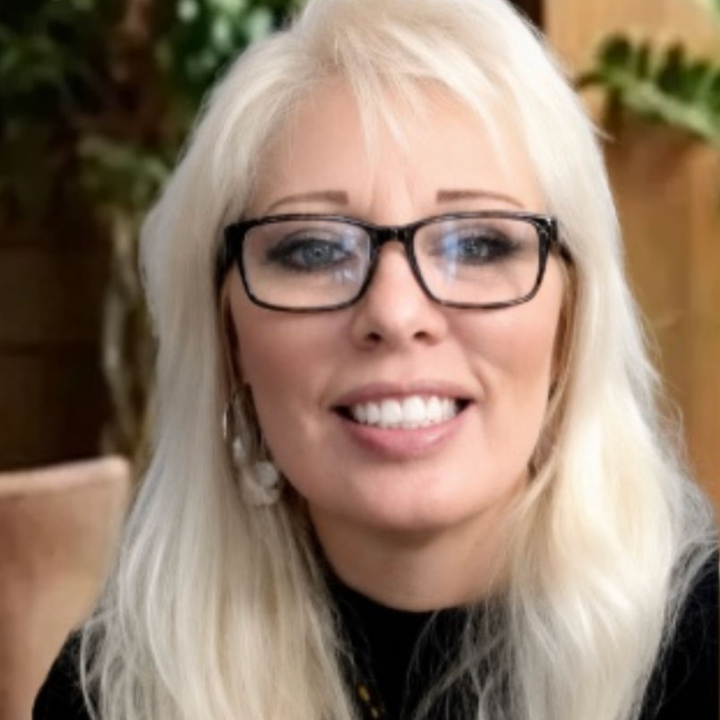 Close-up of a smiling woman with long blonde hair, wearing black-rimmed glasses, earrings, and a black top, sitting in front of a wooden and green outdoor background.