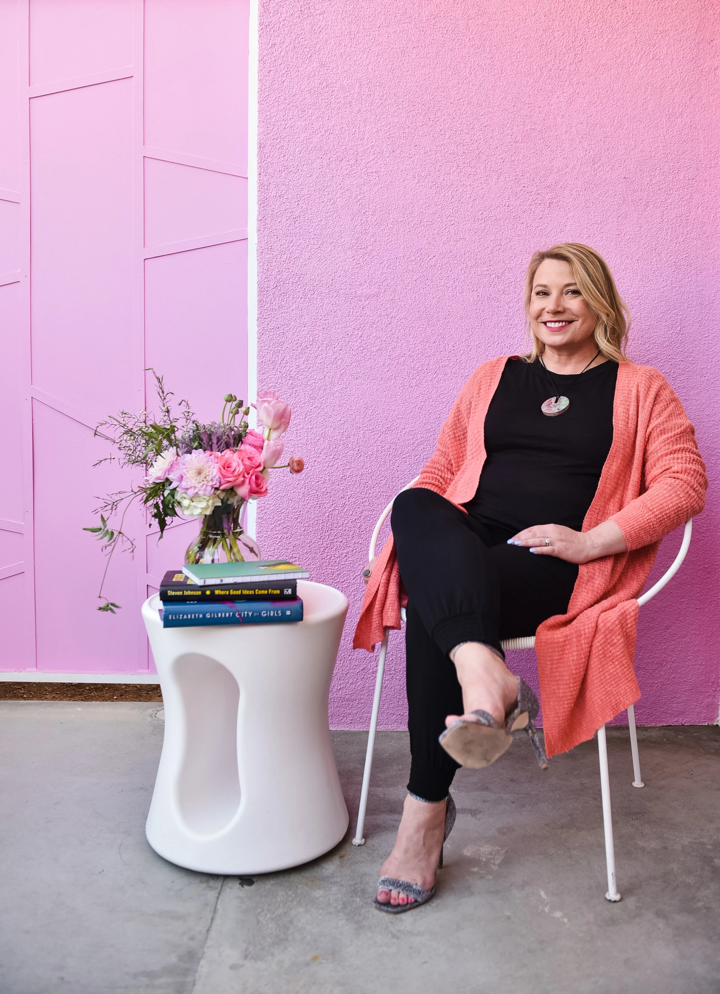 Woman sitting on a white chair, smiling, wearing a black top, black pants, and a pink cardigan. A table with a bouquet of pink and purple flowers and several books is beside her. The background features a pink textured wall and a pink cabinet.