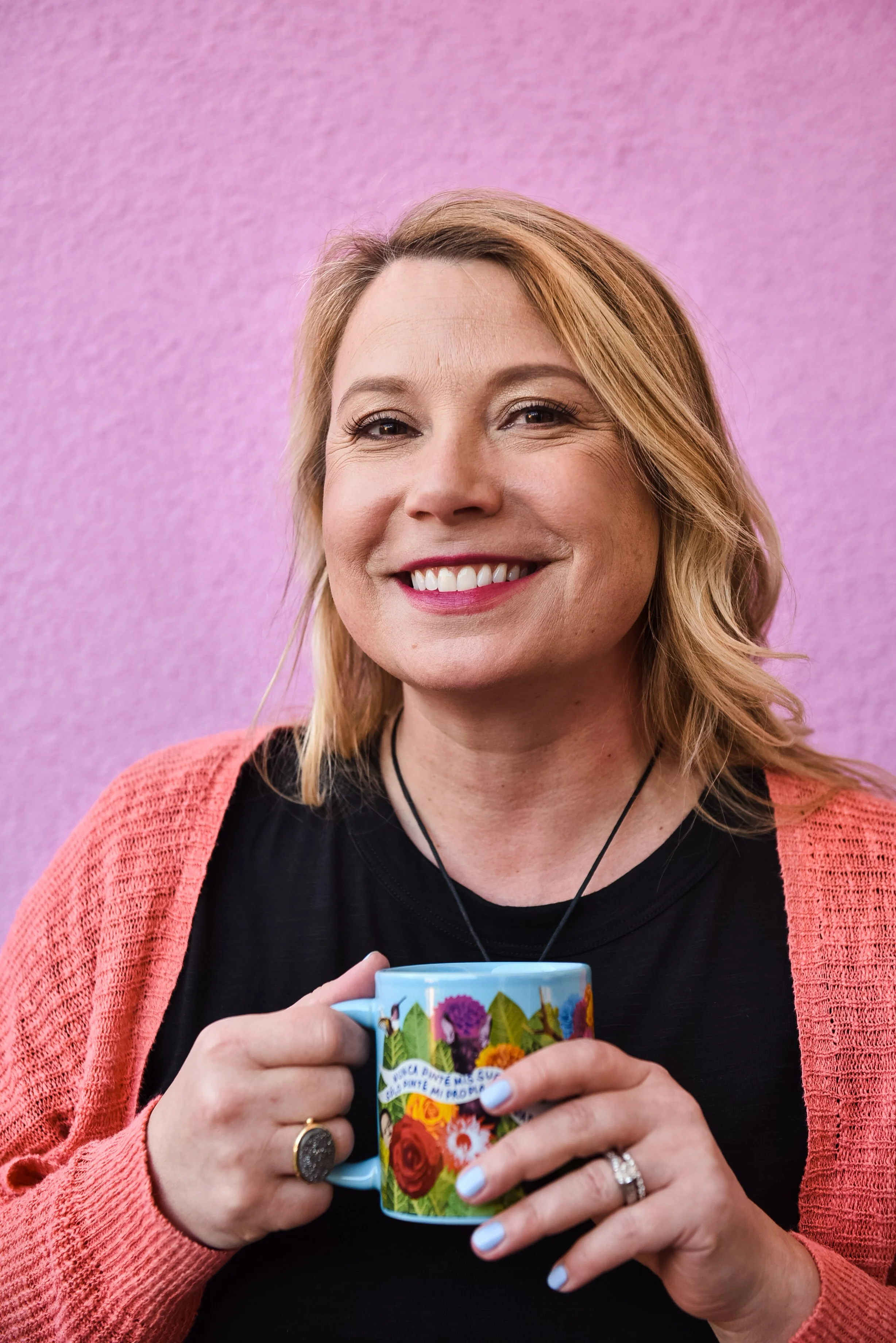 A smiling woman with blonde hair holding a colorful floral mug with text, wearing a black top, coral cardigan, and rings, standing against a pink background.