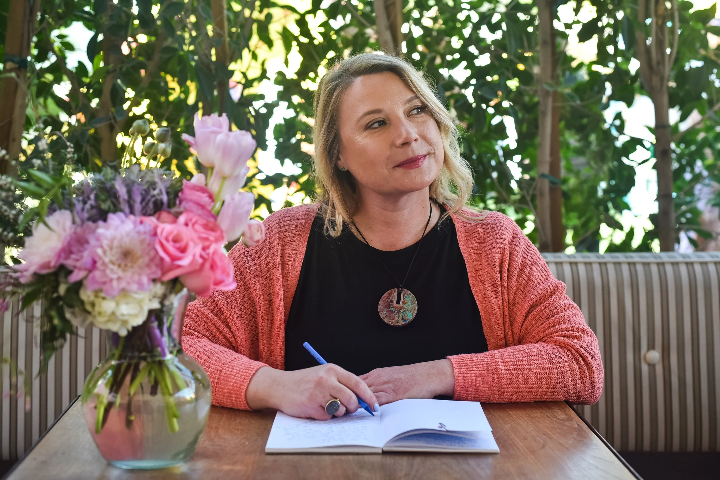 A woman with blonde hair wearing a coral pink cardigan and black shirt sits at a wooden table outdoors, writing on paper with a blue pen. A bouquet of pink, purple, and white flowers in a glass vase is on the table in the foreground. The background features lush green foliage and sunlight filtering through the leaves.